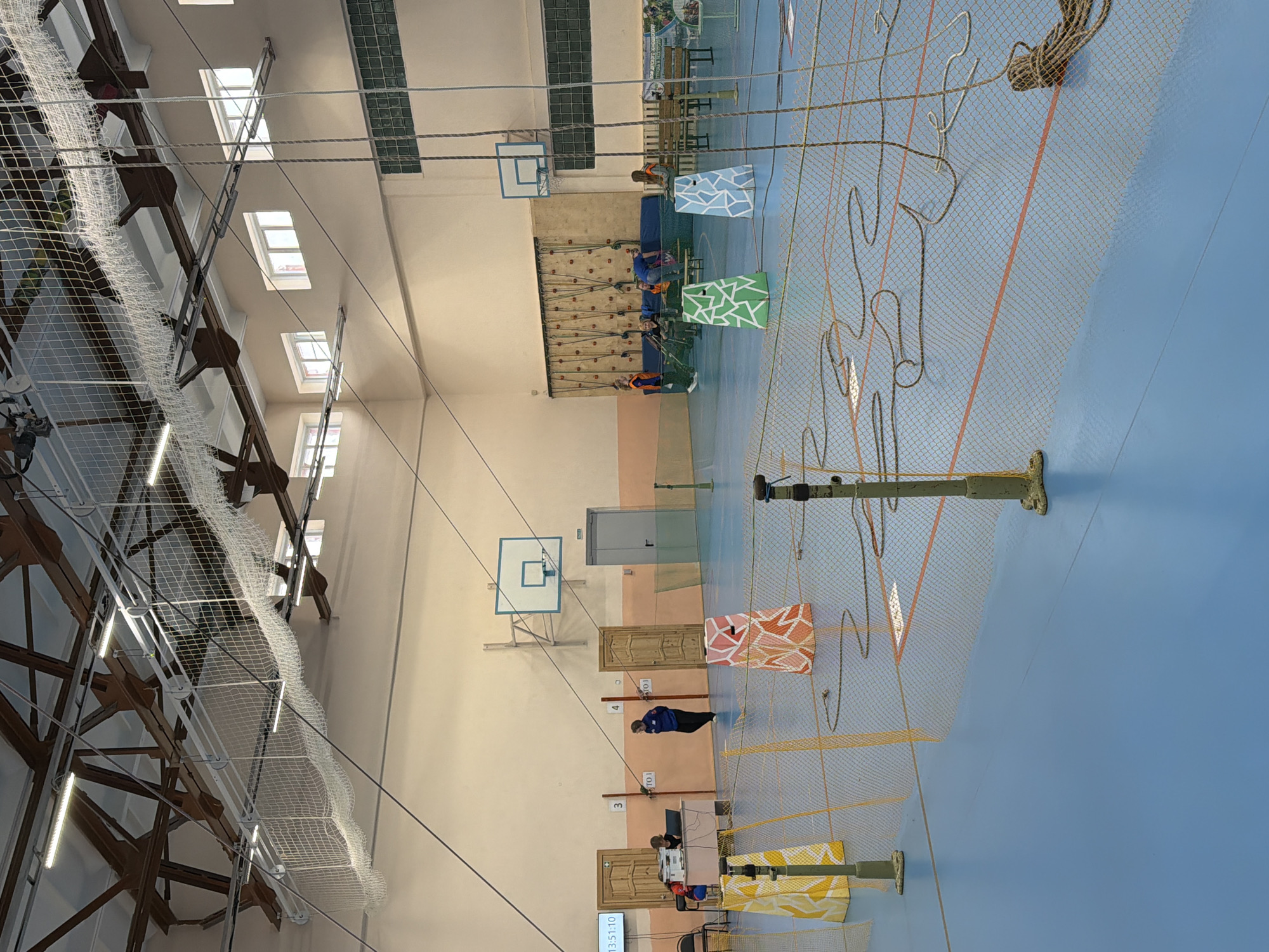 The image shows an indoor sports hall in Berezniki, Russia. The hall has a light blue floor, cream-colored walls, and a high ceiling with exposed metal beams and netting. Natural light enters through multiple windows along one wall. The hall is equipped with basketball hoops and a climbing wall. In the background, several individuals are on the climbing wall. In the foreground, a rope-climbing setup is in use. Ropes hang from above and are connected to colored panels. Some of the panels are marked. People are actively participating in the rope-climbing activity. A clock is visible, showing the time 13:51:10.