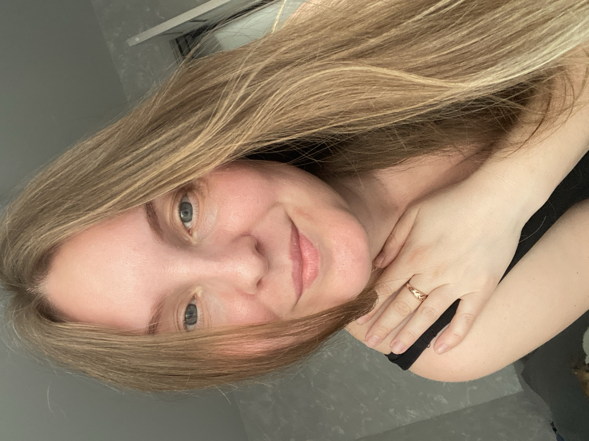 A woman with long blonde hair is photographed in a selfie. She has light skin, blue eyes, and is smiling slightly. She is wearing a black top. Her left hand is visible, and a gold ring is on her ring finger. The background appears to be a room with grey walls. A window is visible in the upper left corner. The photograph is rotated counter-clockwise. There is no indication of the location in the city of Noviy, Russia.