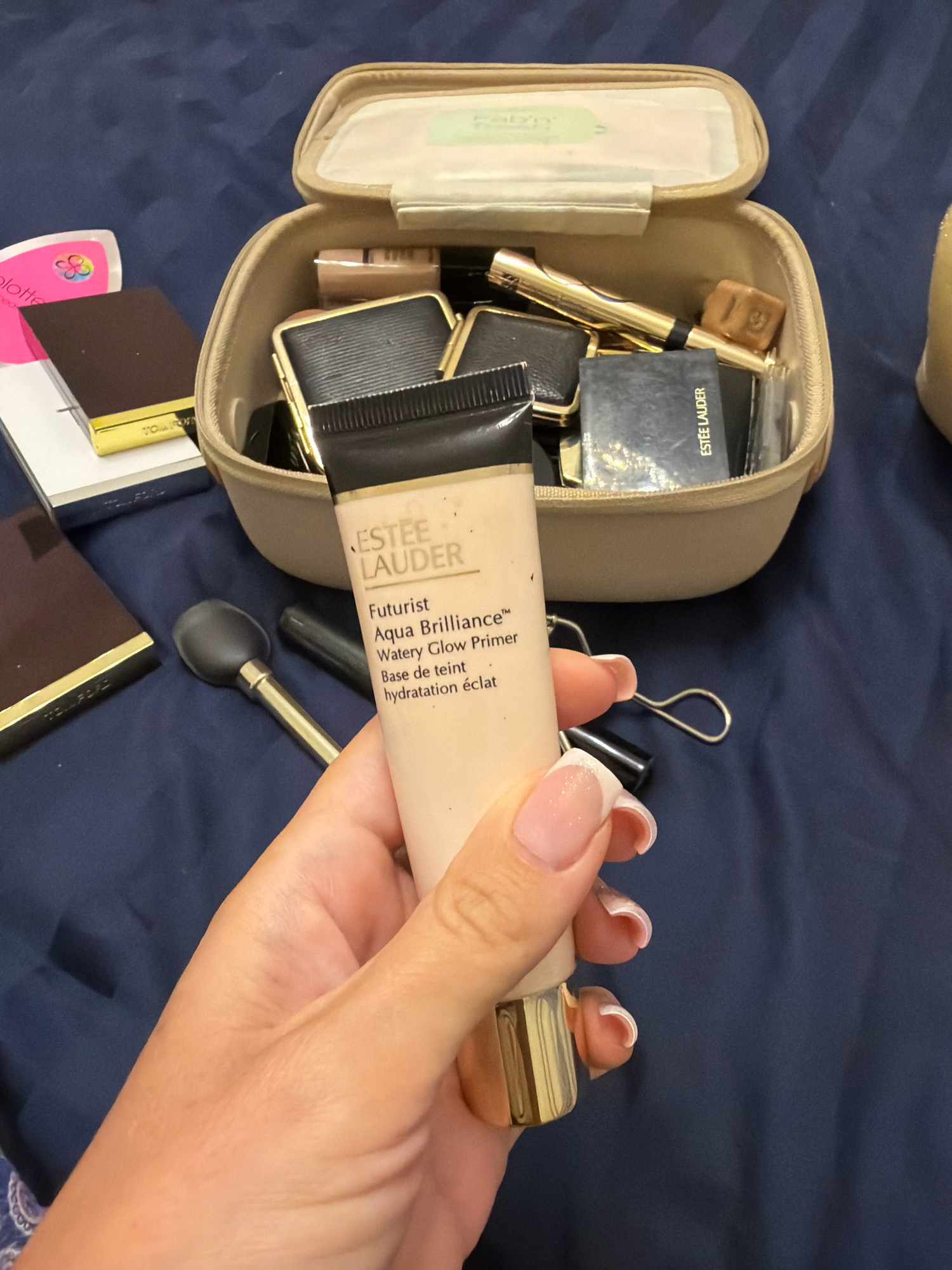 A hand with French manicured nails holds a tube of Estee Lauder Futurist Aqua Brilliance Watery Glow Primer. The tube is pale beige with a black cap and a gold base. In the background, an open tan makeup bag contains various cosmetic items, including what appear to be eyeshadow palettes and a gold lipstick or eyeliner. To the left of the makeup bag are two dark brown and gold compacts, one of which has "TOM FORD" visible. A black makeup brush with a rounded head and gold accents lies on a dark blue fabric surface, which is likely a bedspread or comforter. The scene does not contain any elements that would indicate a specific location in Dubai, United Arab Emirates.