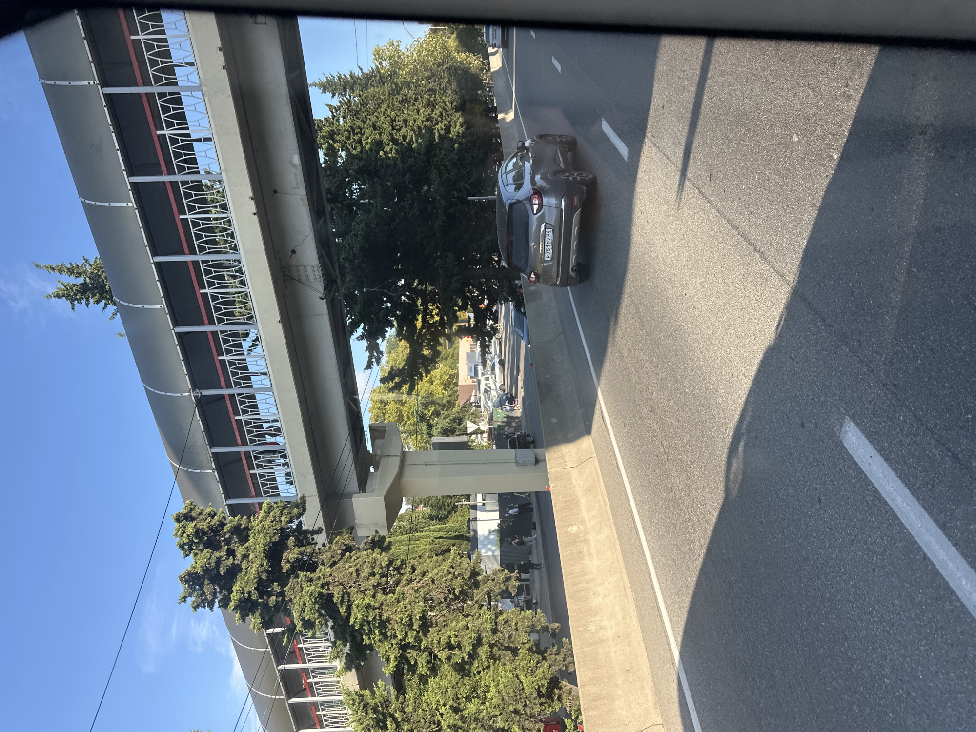 The image shows a view from a moving vehicle on a highway in Sochi, Russia. The highway has a concrete barrier on the left side and a pedestrian bridge overhead. The bridge is gray with a black canopy and white railings. Several evergreen trees are visible along the roadside and under the bridge. On the right side of the highway, a dark gray Renault Captur is driving. Further down the road, other vehicles and buildings can be seen. The sky is clear and blue. The image appears to have been taken in the daytime.
