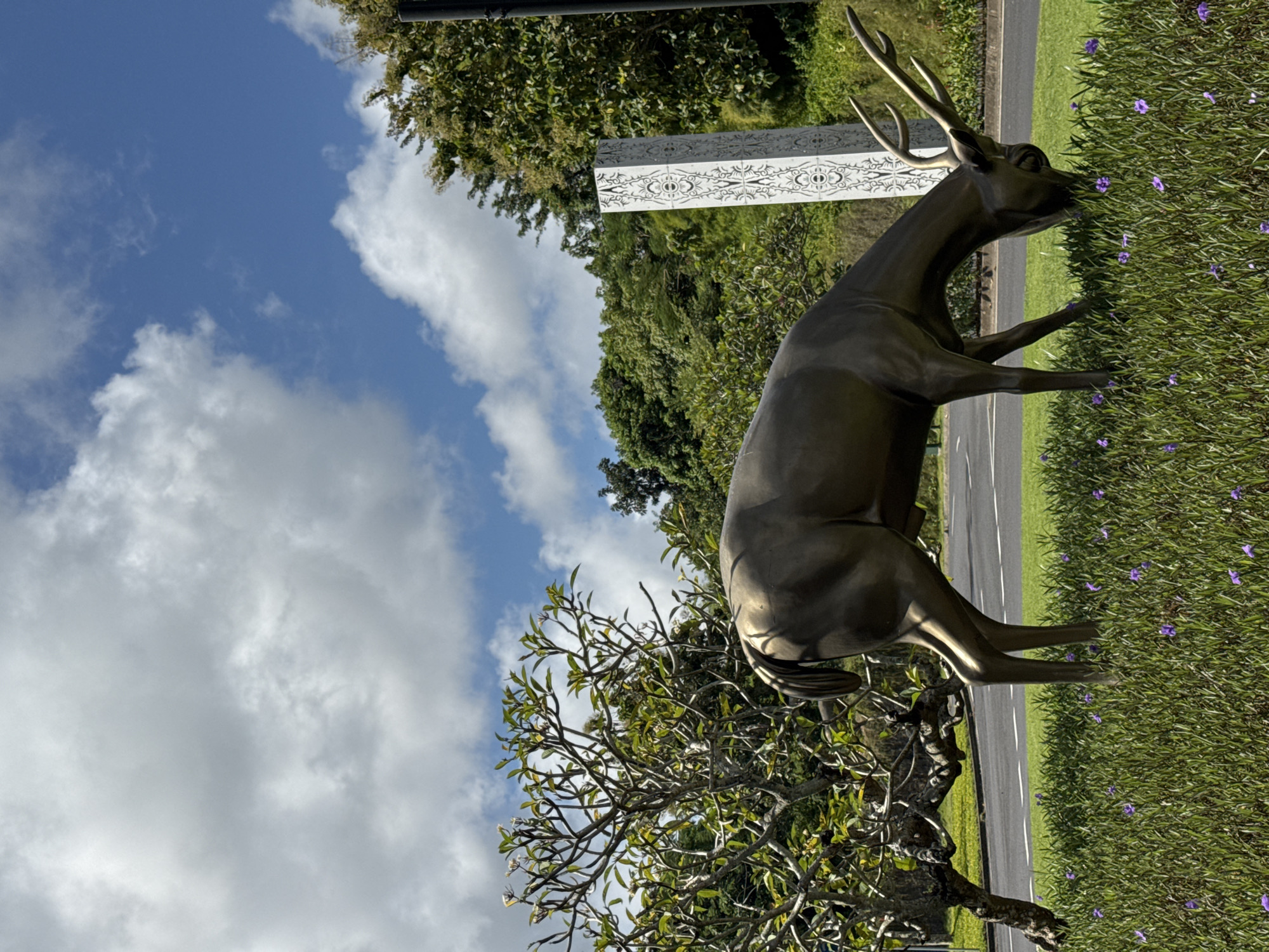A bronze statue of a deer stands on a grassy strip alongside a road. The deer is facing right, appearing to graze among purple flowers. A decorated white concrete block stands behind the deer, partially obscured by foliage. A tree with bare branches is in the foreground. A road runs parallel to the grassy area. The sky is blue with scattered white clouds. The location is Unknown, Unknown.