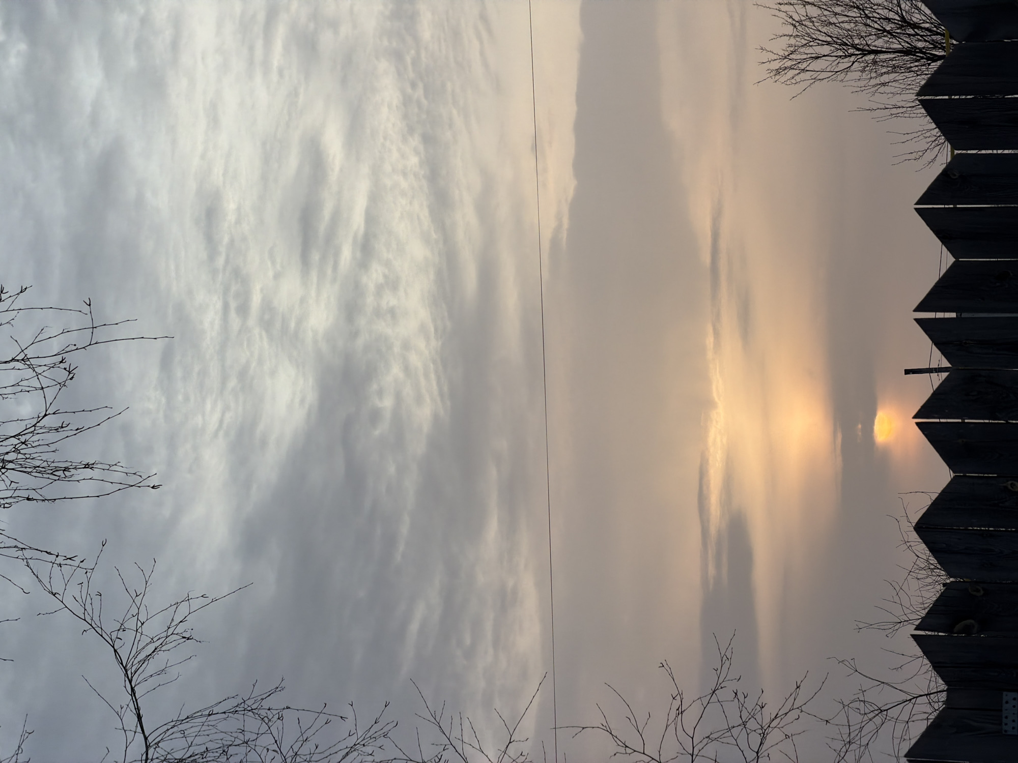 The image captures an outdoor scene at either sunrise or sunset in Chaikovsky, Russia. The sky exhibits a layered appearance, with a dense, textured grey and white cloud cover dominating the upper portion. Below this, the sky transitions into a pale orange-yellow hue, indicative of the sun positioned low on the horizon. The sun itself is partially obscured by clouds, appearing as a bright orange-yellow disc just above the fence line. A single, dark, thin wire, consistent with a power or communication line, stretches horizontally across the middle ground. The foreground features a dark silhouette of a wooden picket fence with sharply pointed vertical slats, occupying the bottom fifth of the frame. Bare, leafless tree branches are silhouetted against the sky, extending from the top-left and mid-left corners, with additional sparse tree branches visible behind the fence on the right side. No individuals or active subjects are present.