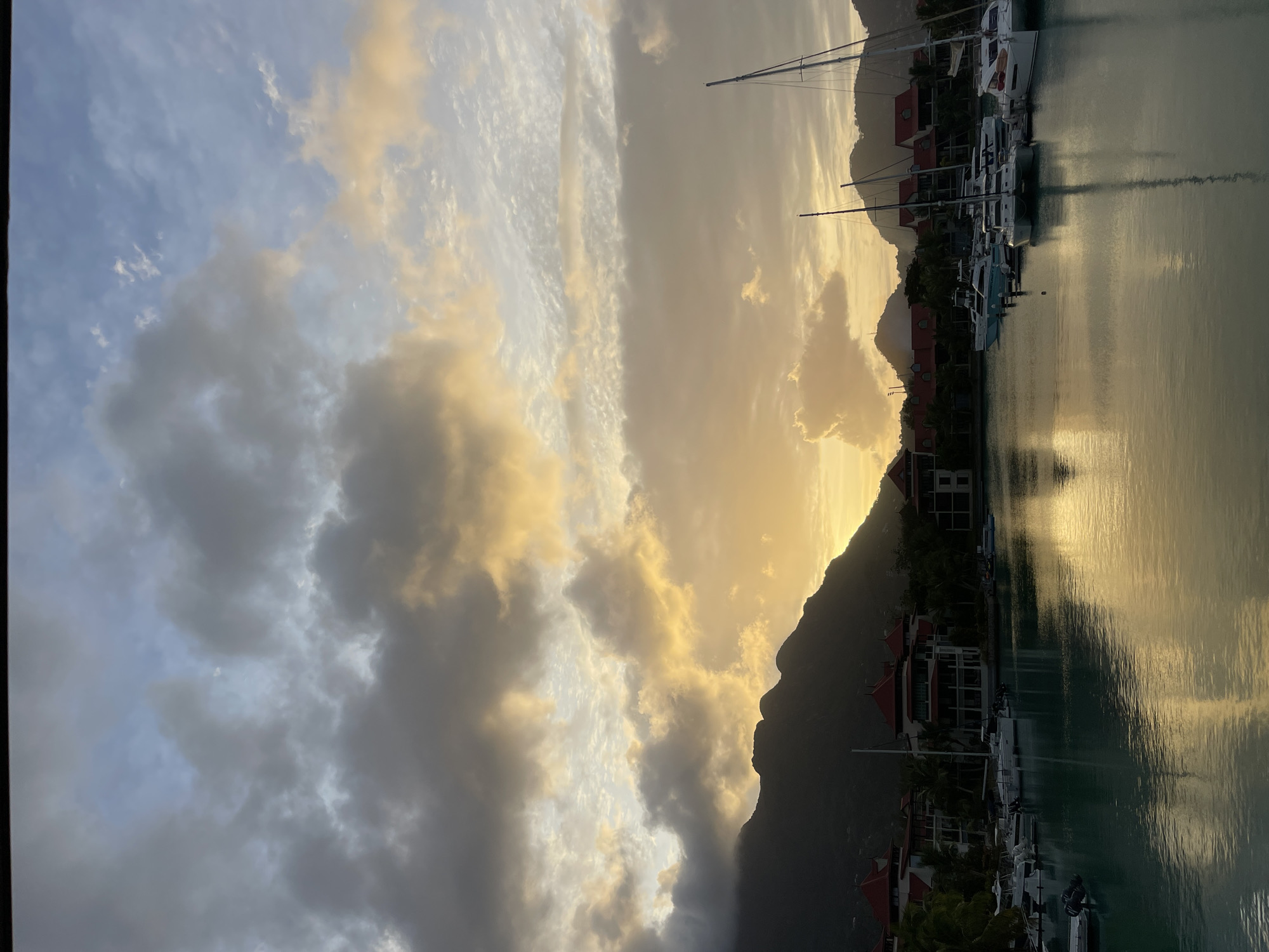 The image depicts a marina in the city of Victoria, Seychelles, during a sunset. The sky is filled with dramatic clouds, some dark and heavy, others illuminated by the setting sun with shades of yellow and orange. Silhouetted mountains form the backdrop, their peaks softened by the twilight.
In the foreground, a body of calm water reflects the colors of the sky and the surrounding structures. Several sailboats and smaller boats are docked in the marina. The boats are modern and appear to be in good condition.
Along the waterfront, there are buildings with red tiled roofs and white walls, characteristic of resort or residential architecture in tropical locations. Lush green trees are interspersed among the buildings. The overall impression is one of tranquility and natural beauty, enhanced by the warm hues of the sunset. There are no people visible in the image. The scene is static, capturing a moment in time.