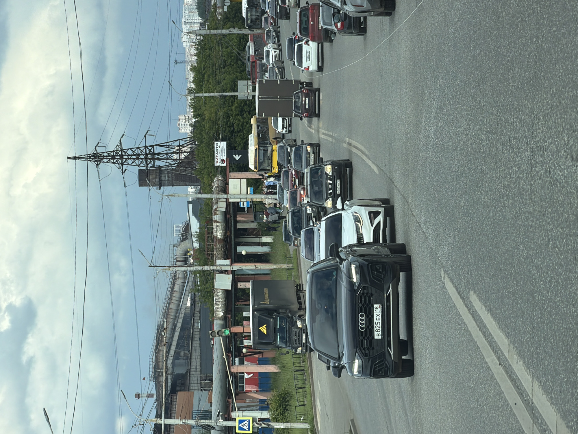 The image shows a traffic scene on a multi-lane road in an urban industrial area under a cloudy sky. In the foreground, a black Audi SUV is prominent, followed by a white sedan. Several other vehicles, including sedans, SUVs, and a yellow bus, are visible in lanes ahead and to the right. On the left side of the road, there is a pedestrian crossing with a yellow diamond-shaped sign. Further back, a dark-colored truck with a yellow logo is stopped at a traffic light, which is green. Industrial buildings with pipes and structures are visible in the background, along with a large electrical pylon. The road surface is asphalt with white lane markings. Trees and smaller buildings are seen in the distance beyond the industrial zone.