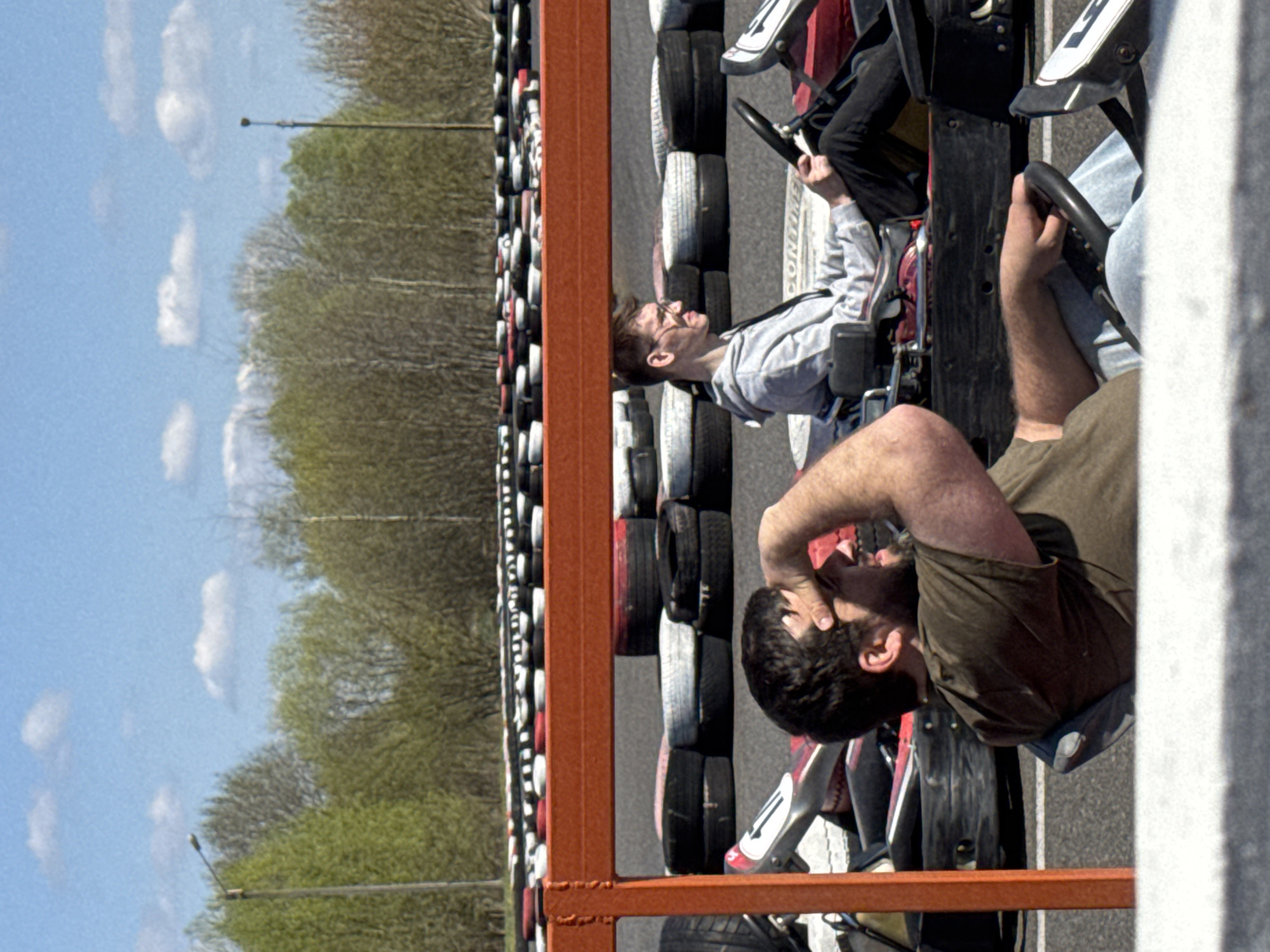 The image depicts a go-kart track in Hrodna, Belarus, with two individuals in go-karts. The go-kart track is bordered by stacks of tires, and a metal barrier separates the track from the surroundings. The sky above Hrodna is visible, showing a clear blue sky with scattered clouds. The individuals in the go-karts are a man in a gray hooded sweatshirt and a man in an olive-green t-shirt. The man in the olive-green t-shirt has his hand on his head and is looking up, while the man in the gray hooded sweatshirt is looking straight ahead.