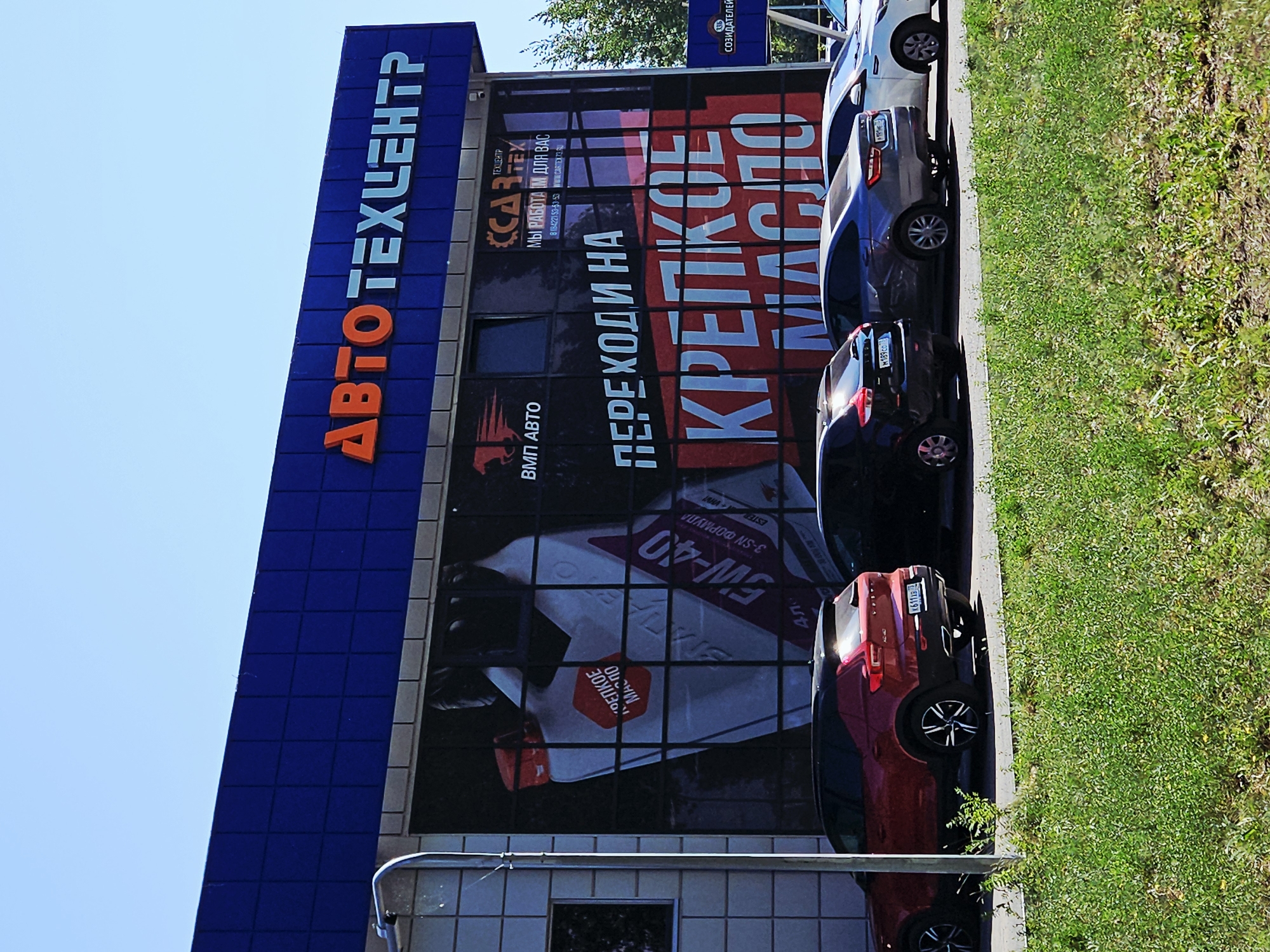The image shows the exterior of an auto service center named "АВТОТЕХЦЕНТР" in Ulyanovsk, Russia. The building has a blue facade with the name of the center displayed in large orange and light blue letters. The large glass windows of the building feature a prominent advertisement for motor oil, with a graphic of a motor oil bottle and the text "ПЕРЕХОДИ НА КРЕПКОЕ МАСЛО" (Switch to strong oil) in large red letters. Several cars are parked in front of the building, including a red Volvo XC40, a dark-colored sedan, and a white sedan. The weather appears to be sunny, with a clear blue sky. The foreground is filled with lush green grass.