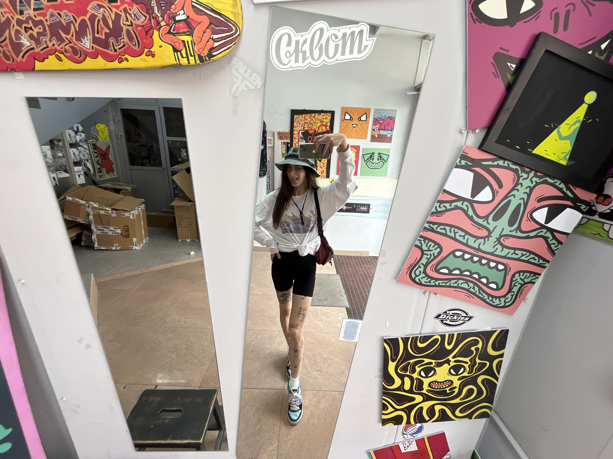 The image depicts a young woman taking a selfie in a full-length mirror, likely within an urban studio or retail space. The woman is wearing a white long-sleeved shirt, black shorts, a green bucket hat, and brightly colored sneakers. She has several tattoos on her legs. In the background, reflected in the mirror, are stacks of cardboard boxes, a doorway, and various pieces of artwork and skateboards adorning the walls. The overall aesthetic suggests a creative or retail environment, possibly a skate shop or an artist's studio. The specific location in Krasnoyarsk, Russia, cannot be determined from the visual information provided.