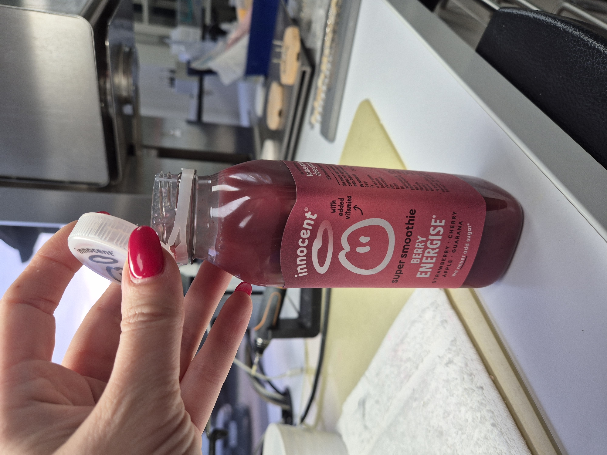 A person with red nail polish is opening a bottle of Innocent Super Smoothie Berry Energise. The bottle contains a dark red liquid. The background is blurred, showing a countertop and equipment. The location appears to be indoors, possibly a kitchen or food preparation area. The city and country cannot be inferred from the image.