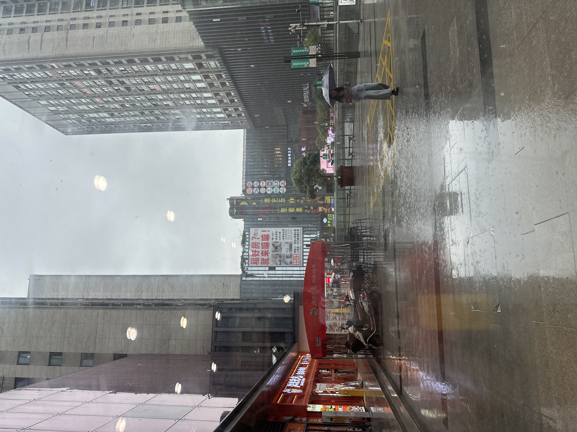 The image depicts an urban street scene in Chongqing High-tech Zone, China, on a rainy, overcast day. The photograph is taken from an interior vantage point through a glass window, evidenced by distinct circular light reflections superimposed on the external environment.
The foreground is a wet, tiled pavement, reflecting ambient light and surrounding structures, with visible raindrops impacting the surface. Yellow cross-hatch road markings are present on the ground in the mid-ground.
To the immediate left, a building with reddish-pink exterior panels features a brightly lit ground-floor shop with prominent red signage, displaying Chinese characters including "麻辣烫" (Malatang) and "龙坎" (Long Kan), indicating a food establishment. Adjacent to this shop, a red awning extends over an outdoor seating area furnished with dark metal chairs and tables. A white scooter is parked beneath this awning.
In the mid-ground, a lone individual, appearing female, walks from right to left across the wet pavement, holding a black umbrella. She is wearing light-colored trousers and a top.
The background is characterized by numerous tall, modern, grey multi-story buildings. One prominent building displays a large, illuminated sign with Chinese text that includes "租好房?就来嘛!" (Rent a good house? Come on!), accompanied by illustrative images of interiors. Another building to the left exhibits a vertical array of diverse, colorful circular commercial logos. Sparse trees in planters are visible between the structures and in the central plaza area. The overall scene conveys a commercial district experiencing rainfall.