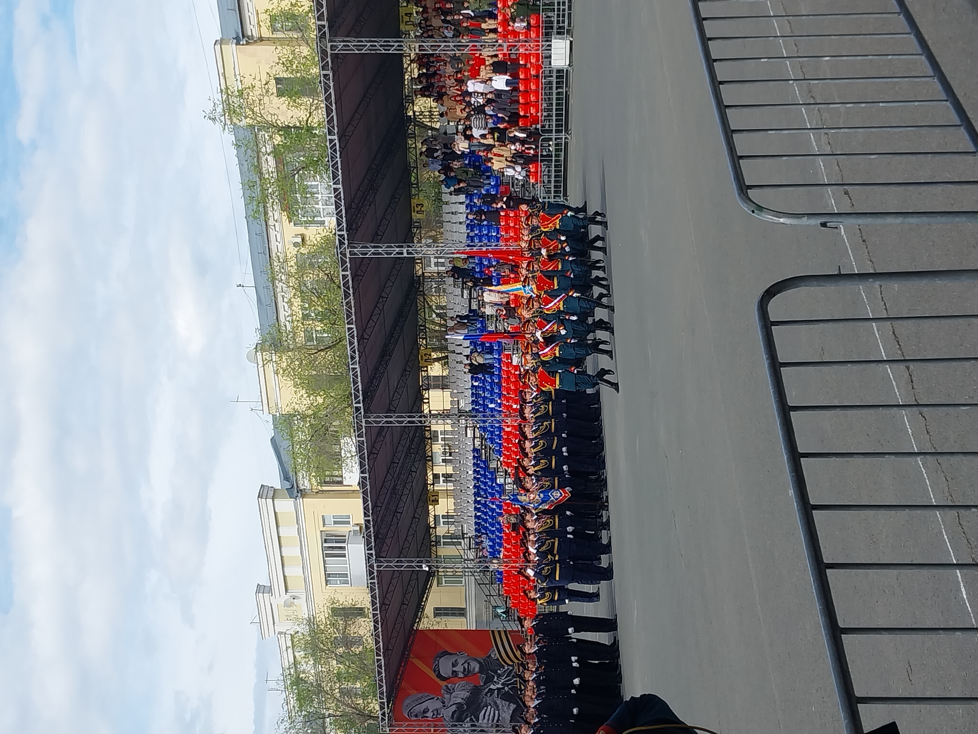 The image depicts a military parade in Orenburg, Russia. A column of soldiers marches along a street lined with spectators in tiered seating. A large banner with portraits is visible at the end of the parade route. The architecture of nearby buildings is typical of urban Russia. Metal barricades line the side of the street. The sky is partly cloudy.