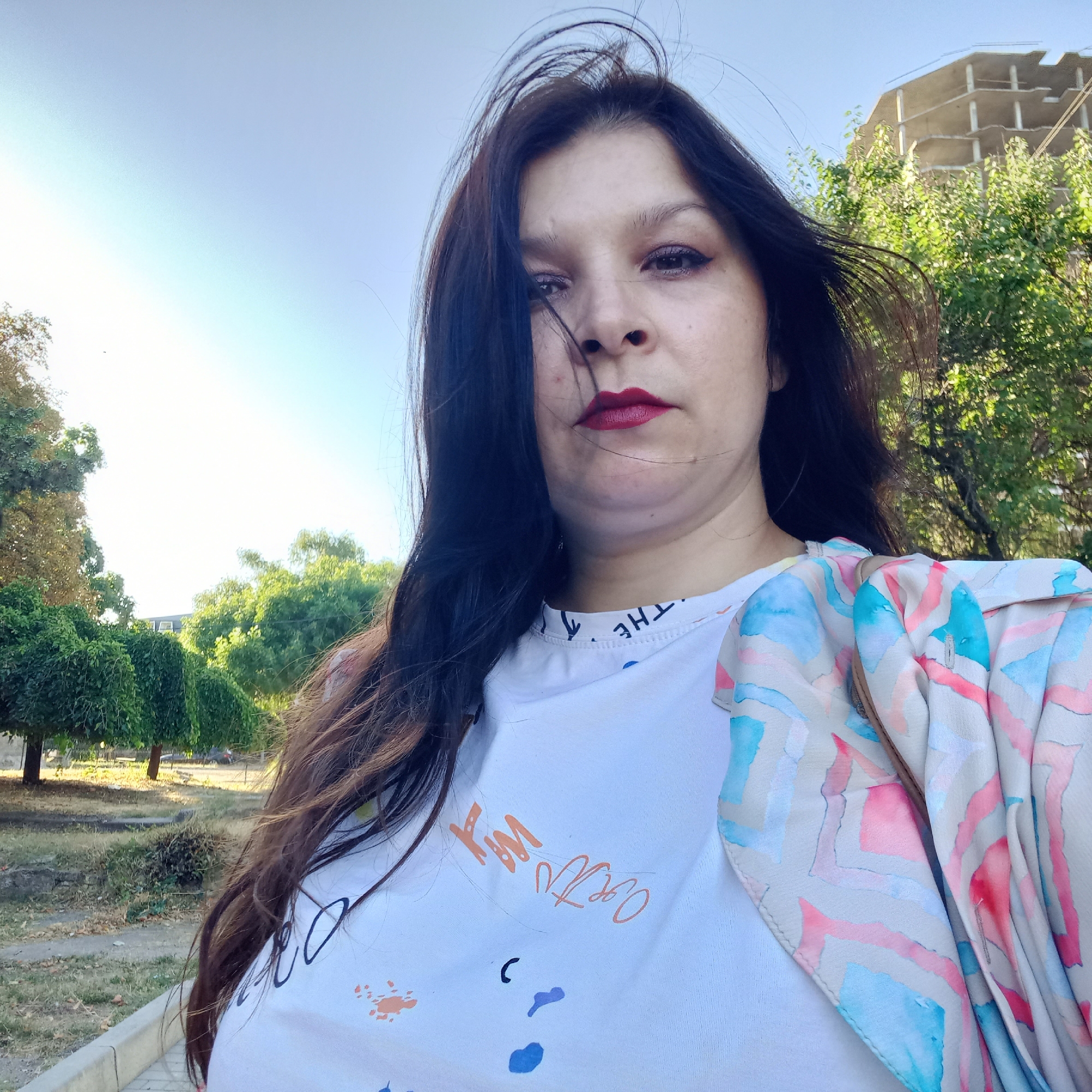 A woman with long dark hair and red lipstick poses for a selfie outdoors. She is wearing a white t-shirt with an abstract design and a light-colored jacket with a watercolor-like pattern of pink, blue, and beige shapes. Behind her, trees and an unfinished building are visible under a clear sky. The location is Kropyvnytskyi, Ukraine.