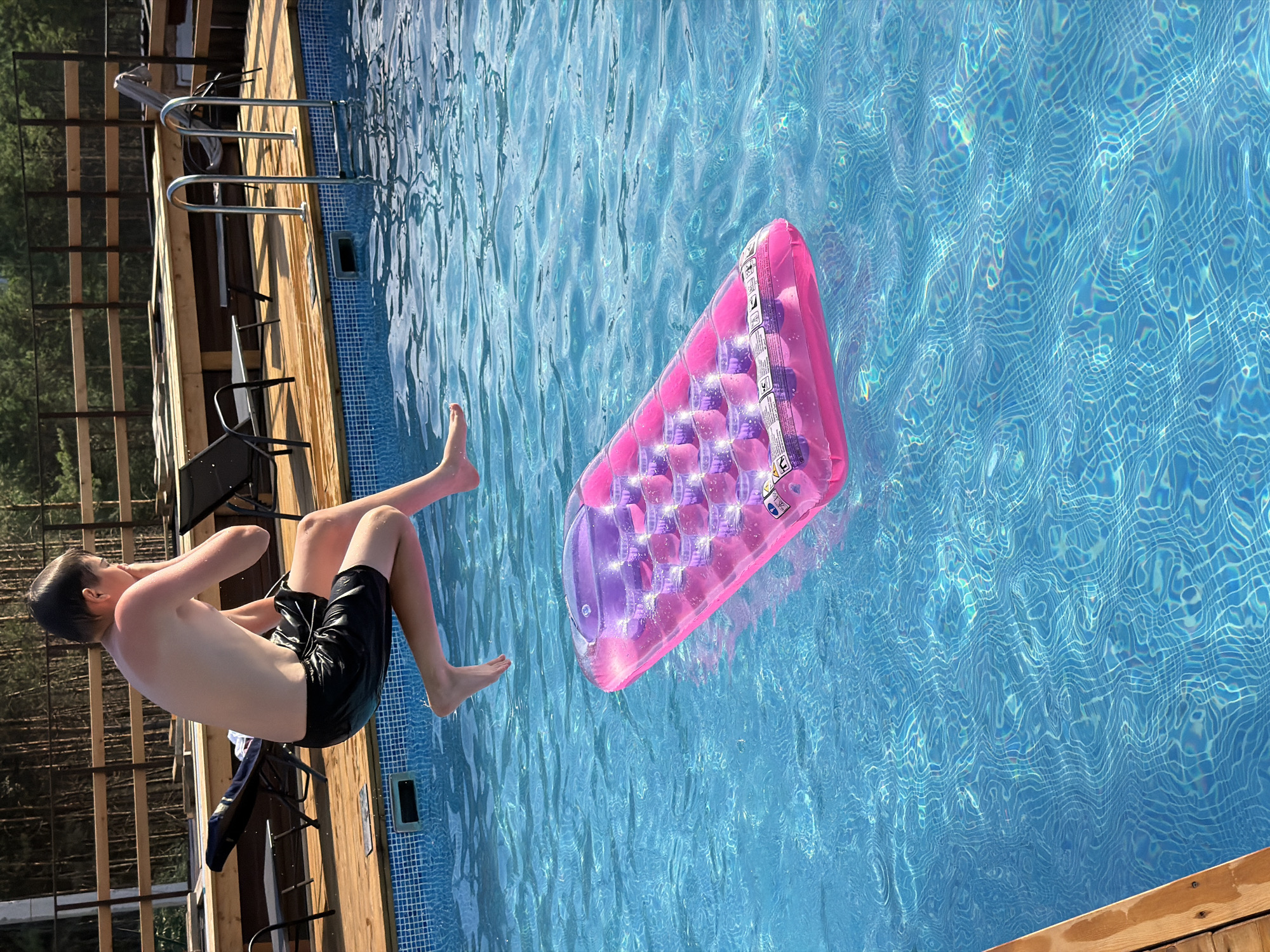 A young boy is captured mid-air, in the process of jumping into a bright blue swimming pool. He is wearing black swim trunks. His body is contorted as he enters the water, with his arms bent and his legs extended. To his right, a pink and purple inflatable pool raft floats on the surface of the water. The pool is surrounded by a wooden deck, and a wooden fence with metal railings is visible behind the boy. Trees can be seen in the background, suggesting an outdoor, natural setting. The water is clear and reflects the sunlight, creating shimmering patterns on the surface. The lighting indicates it is daytime, likely sunny, given the bright reflections and shadows. The scene evokes a sense of summer fun and recreation. There is no visible text in the image. The location context is Elekmonar, Russia.