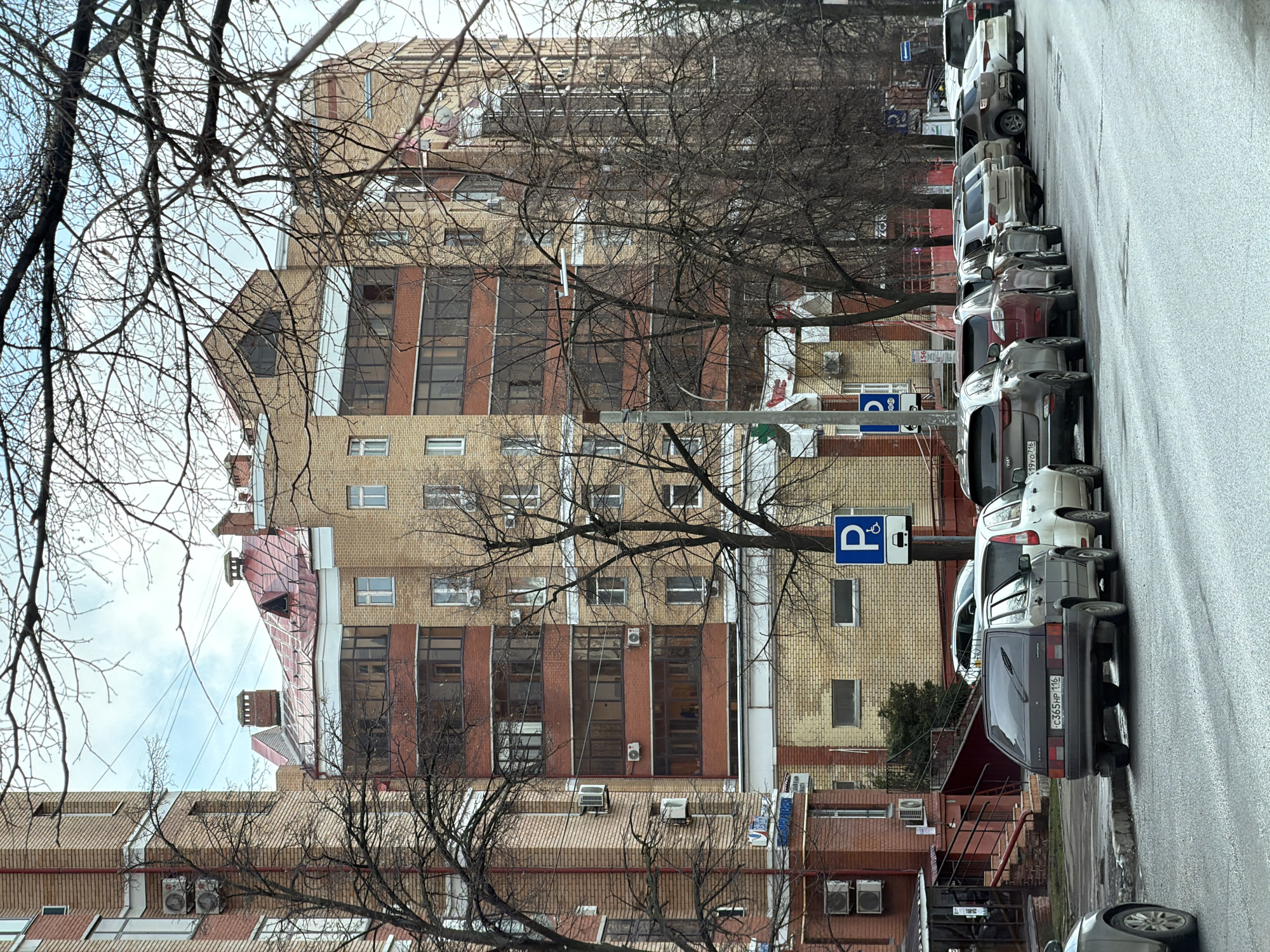The image depicts a street in Kazan, Russia. It shows a multi-story brick building with visible windows, some with red brick framing. Several bare trees are in the foreground, partially obscuring the view of the building. A row of parked cars lines the right side of the street. A blue and white parking sign is visible. The street surface appears to be wet.