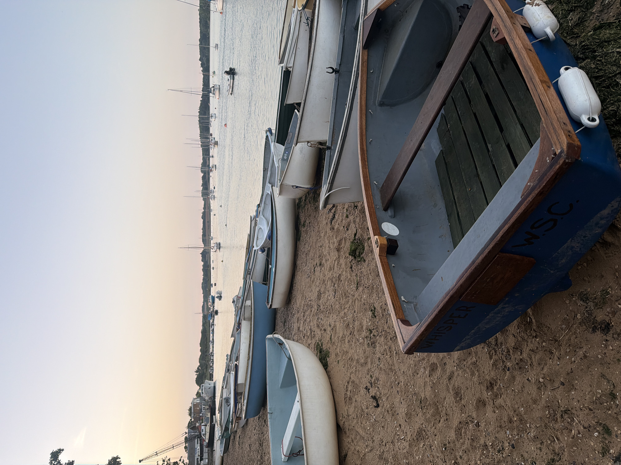 The image shows a row of small boats resting on a sandy beach. The boats are of varying sizes and colors, with some appearing to be made of wood and others of fiberglass. In the background, there's a body of water dotted with sailboats and other vessels. The sky is a gradient of light hues, suggesting either sunrise or sunset. The location is likely East Suffolk, United Kingdom, given the coastal setting and the presence of moored sailboats.