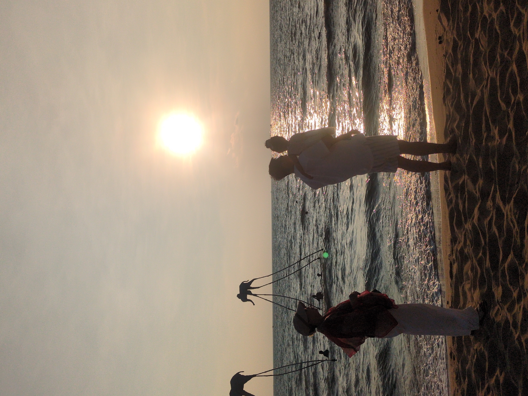 A beach scene at sunset in Phú Quốc, Vietnam, shows a person in white clothing, possibly a local vendor, with two contraptions holding up what appear to be kites shaped like kangaroos. Near the ocean, a couple stands embracing, silhouetted against the setting sun. The tide is coming in, reflecting the light.