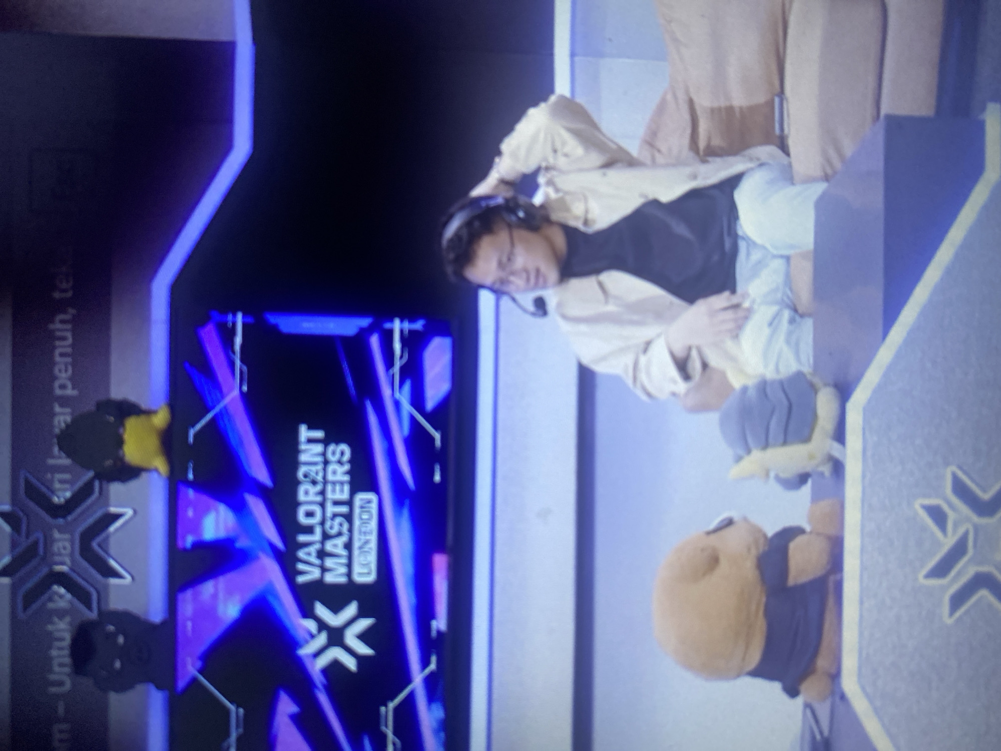 The image captures a man wearing a black t-shirt, light-colored jacket, glasses, and a headset, seated on a sofa. He is looking to his left with his right hand touching the back of his head. In front of him, on a blue table, are two plush toys: one resembling a brown dog wearing a dark shirt, and another that is blue and yellow. Behind the man, a large screen displays the "VALORANT MASTERS LONDON" logo. Above this logo, text in Indonesian, "m - Untuk keluar dari layar penuh, tekan...", is visible. The scene appears to be set indoors, likely in a studio or event space, possibly related to gaming or esports. There is no visible indication of the city of Lhokseumawe, Indonesia within the image itself.