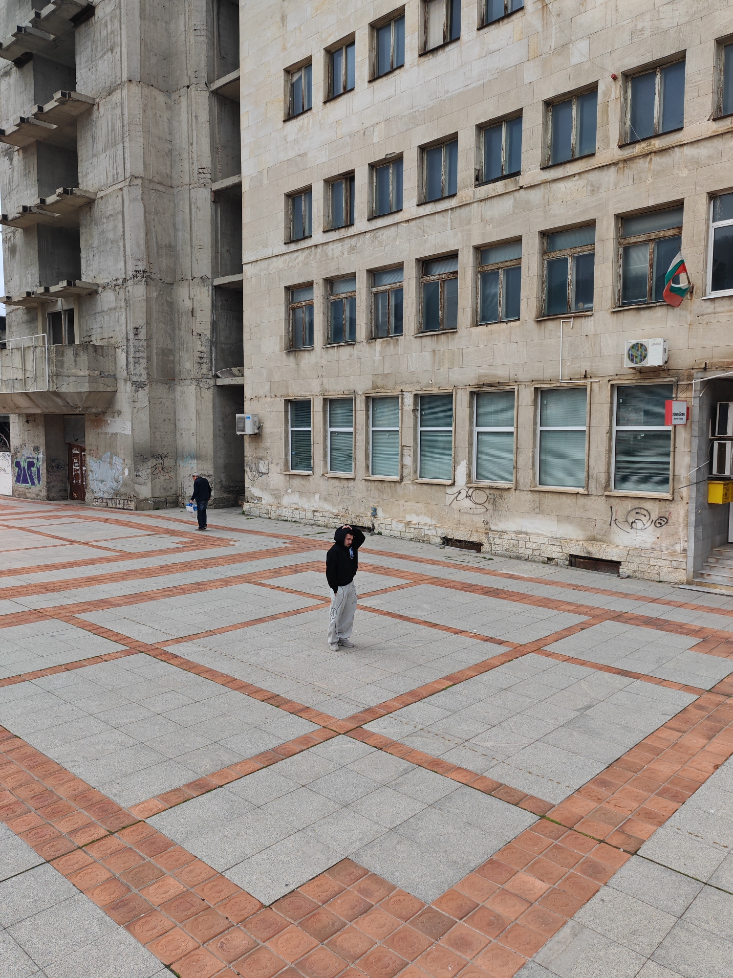 The scene depicts an urban public square in Shumen, Bulgaria, under an overcast sky. The ground is paved with a grid pattern of light grey square tiles and reddish-brown terracotta-style tiles. Two male subjects are present. In the mid-ground, a male wearing a black hooded sweatshirt and light grey sweatpants stands, his left hand positioned near his head. Further to the left and rear, a second male, dressed in dark attire, walks away from the camera carrying a light-colored container in his left hand. No interaction between the two individuals is observed. The background features two buildings. To the right, a multi-story building with a light-colored facade displays multiple windows, an exterior air conditioning unit, a red "MoneyGram" sign, and a yellow utility box near a ground-level entrance. A Bulgarian national flag is affixed to the building's facade on the second floor. To the left, a raw, unfinished concrete structure with exposed slabs and columns exhibits graffiti on its lower walls.