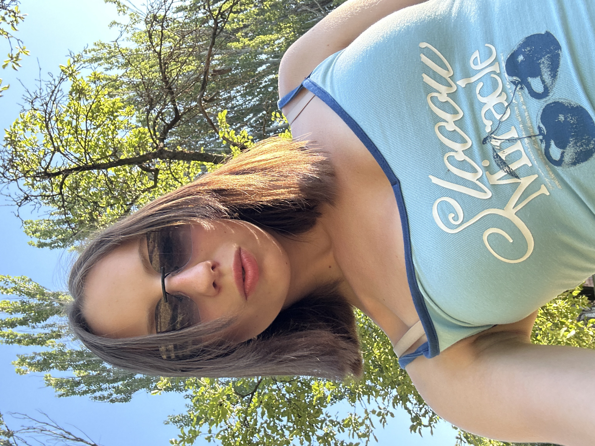 This media file is a selfie taken outdoors on a sunny day, likely in Kryvyi Rih, Ukraine. The subject is a young woman with brown hair and sunglasses, wearing a light blue tank top with a dark blue trim. The tank top has white lettering that appears to read "Slow Miracle" and an image of two cherries. The woman is looking up towards the camera, with a slightly parted mouth. Her pose is casual and relaxed, suggesting she is enjoying the outdoors. The background consists of green trees and a clear blue sky, indicating it is daytime and the weather is pleasant. The low angle of the shot emphasizes the bright sunlight filtering through the leaves. The overall mood is one of carefree enjoyment and summer vibes.