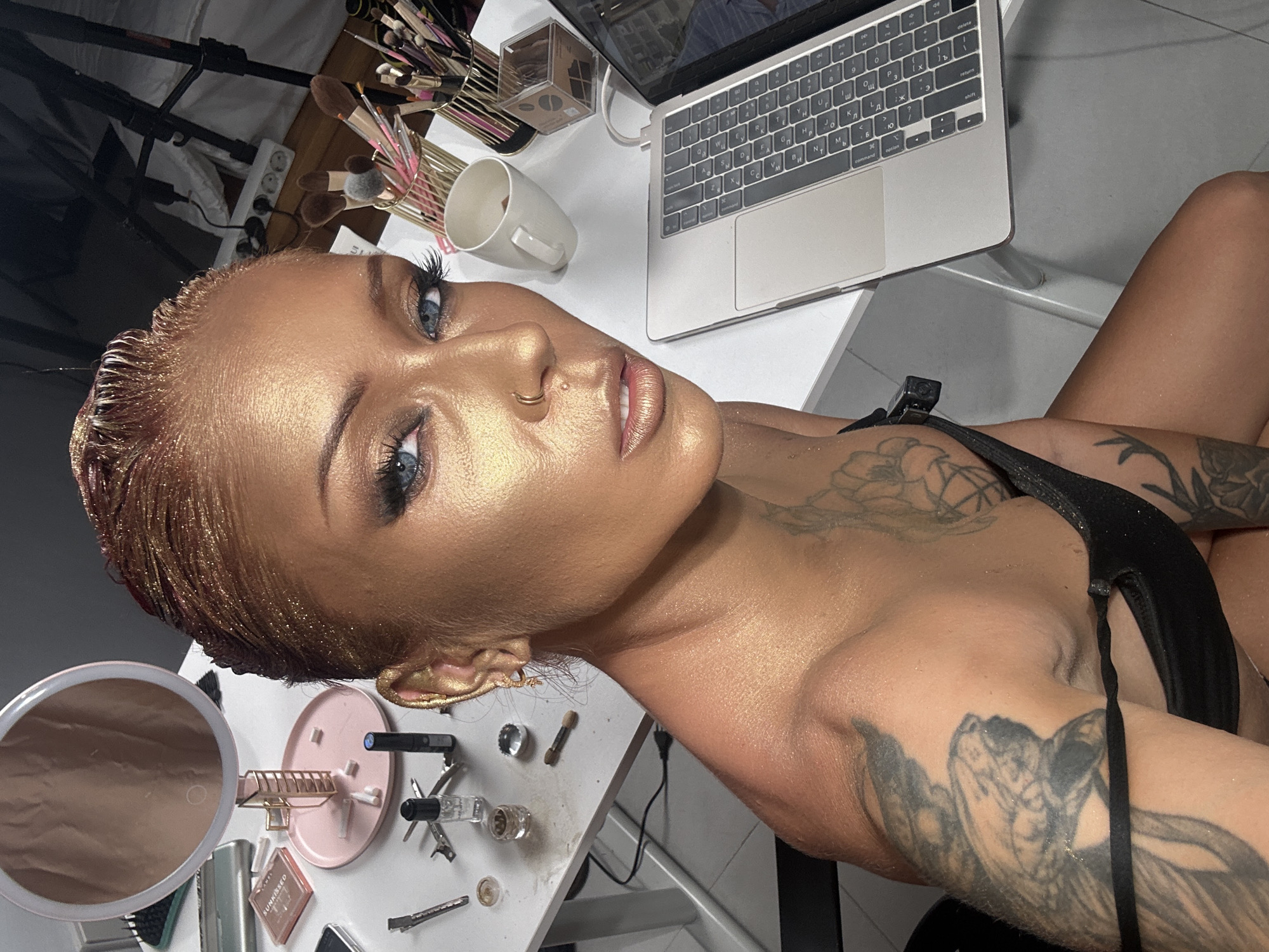 An image, rotated 90 degrees counter-clockwise, features a woman with striking, shimmering golden makeup and hair, looking directly at the viewer with piercing blue eyes. Her skin, neck, and shoulders are covered in a radiant gold glitter, which also highlights her short, styled hair. She has a small gold hoop nose piercing and her lips are painted with a glossy, pinkish hue. Her eyelashes are long and dark. Visible tattoos adorn her arms and shoulders, with intricate designs that include what appears to be floral patterns and possibly an angel-like figure. She is wearing a black top or swimwear.
The woman is positioned in an indoor setting, likely a makeup station or vanity. A white desk or surface occupies the background, cluttered with various items. To her left (top in the rotated image) are several holders filled with numerous makeup brushes. A white ceramic mug is also on the desk. An open silver MacBook laptop sits prominently on the desk, its keyboard visible, showing both English and Cyrillic characters. Other visible items on the desk include a circular light-up makeup mirror, a pink circular tray holding smaller items, various makeup products, applicators, hair clips, and a power strip plugged into an outlet. One makeup compact clearly displays the text "SUNKISSED". The overall impression is one of a glamorous and meticulously prepared look, likely for a photoshoot or event in North Kuta, Indonesia.