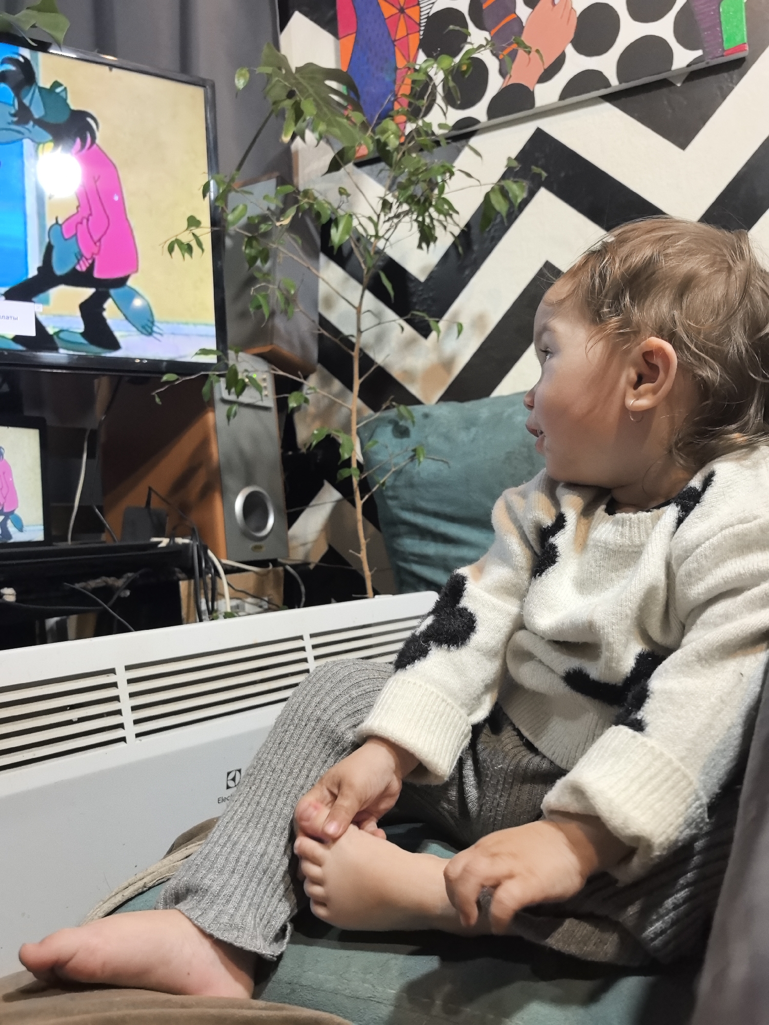A toddler with light brown, curly hair, wearing a white sweater with black cow spots and gray ribbed leggings, sits on a soft surface. The toddler is barefoot, holding their foot with one hand and holding the other hand. The toddler looks to their right. Behind the toddler is a green pillow, a television playing a cartoon, a tall indoor plant, a speaker, and a black and white geometric patterned wall hanging. A white radiator sits in front of the television. The location is implied to be Noviy, Russia.