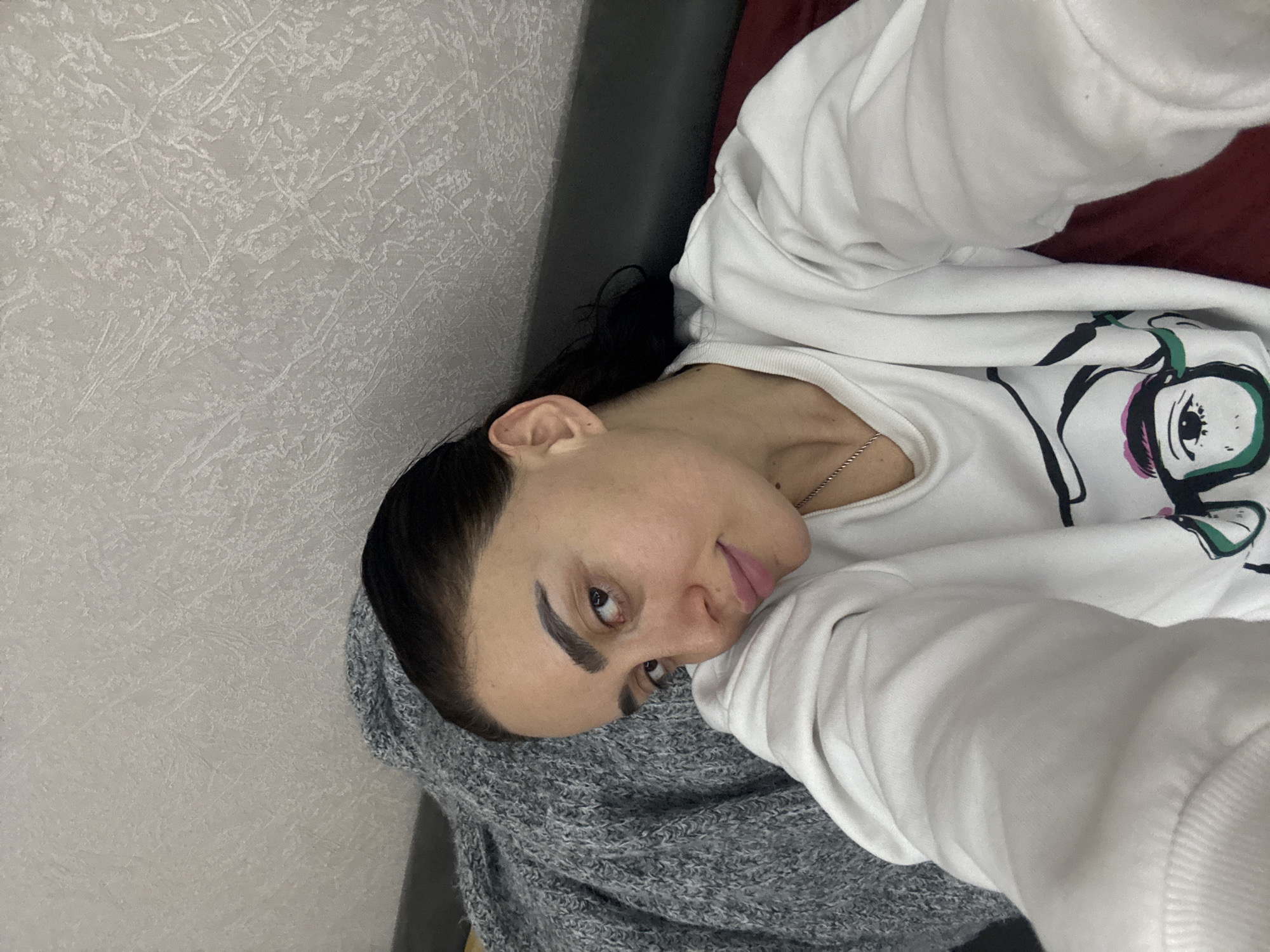 A woman with dark hair is laying down, facing the camera at an angle, in what appears to be an interior setting. She is wearing a white sweatshirt with a graphic design on the front, and a gray knitted garment partially covers her. A small silver necklace is visible around her neck. The background includes a gray headboard of a bed or couch, and a textured white wall. A maroon color is seen above the headboard. The precise location is not apparent from the image, therefore it cannot be determined if the image was taken in Tbilisi, Georgia, or any other location.