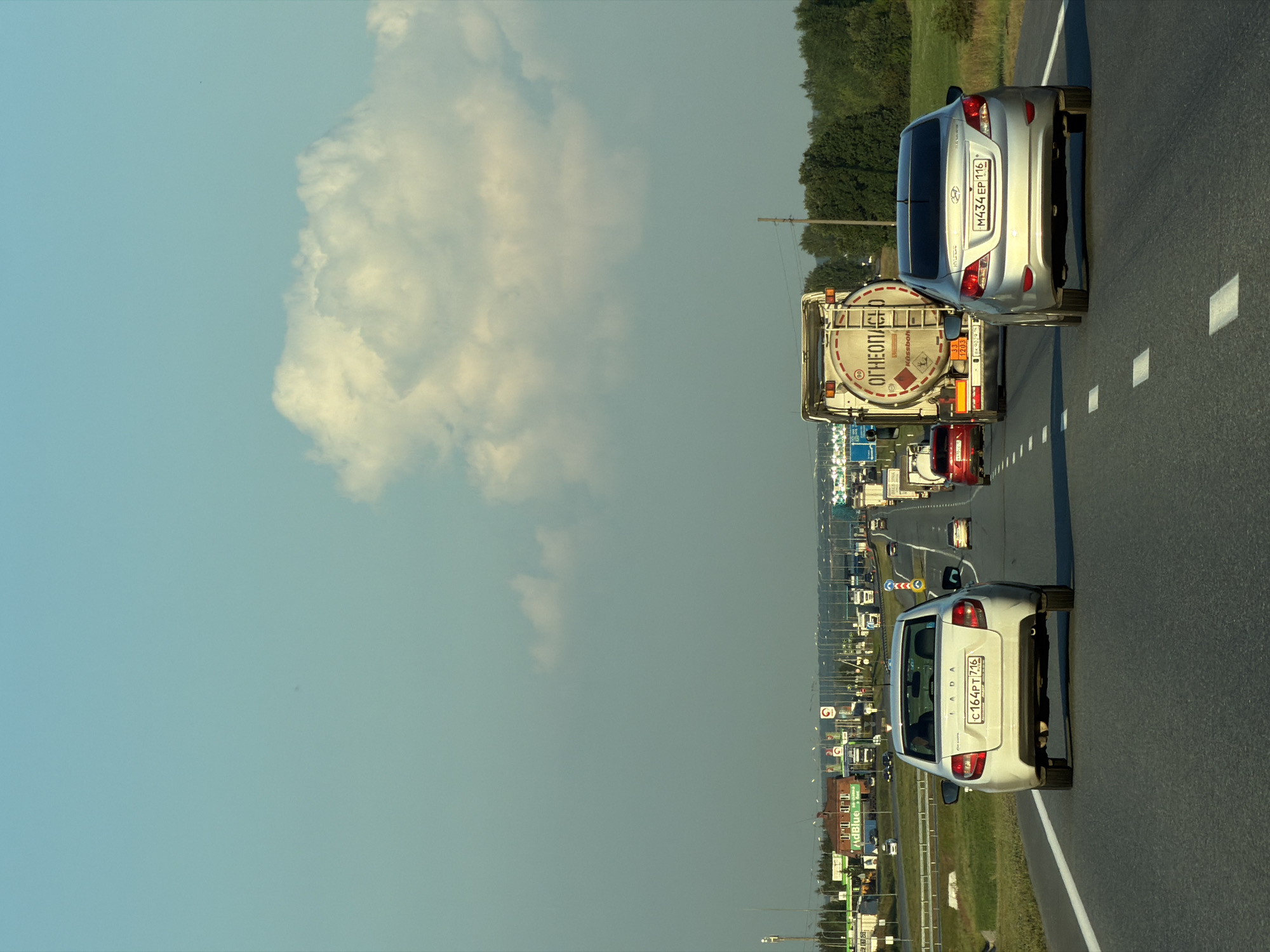 This is a daytime, eye-level shot of a multi-lane highway in the city of Aryshkhazda, Russia. The road is paved asphalt with white dashed lines marking the lanes. The sky is a pale blue with some scattered white clouds. On the right side of the road, there is a dense line of green trees. On the left side, there are some buildings, gas stations, and overhead signs.
In the foreground, a white Lada sedan is driving in the right lane, its rear facing the camera. It has a license plate reading "C164PT 716". To its right, a silver Hyundai sedan is in the left lane, also with its rear facing the camera. Its license plate reads "M434EP 116".
Further down the highway, a large tanker truck is in the left lane. The truck is white and has a large cylindrical tank with the Russian word "ОГНЕОПАСНО" (flammable) printed on it. Other vehicles, including more trucks and cars, are visible in the distance, heading in the same direction. The overall scene depicts moderate highway traffic under a clear sky.