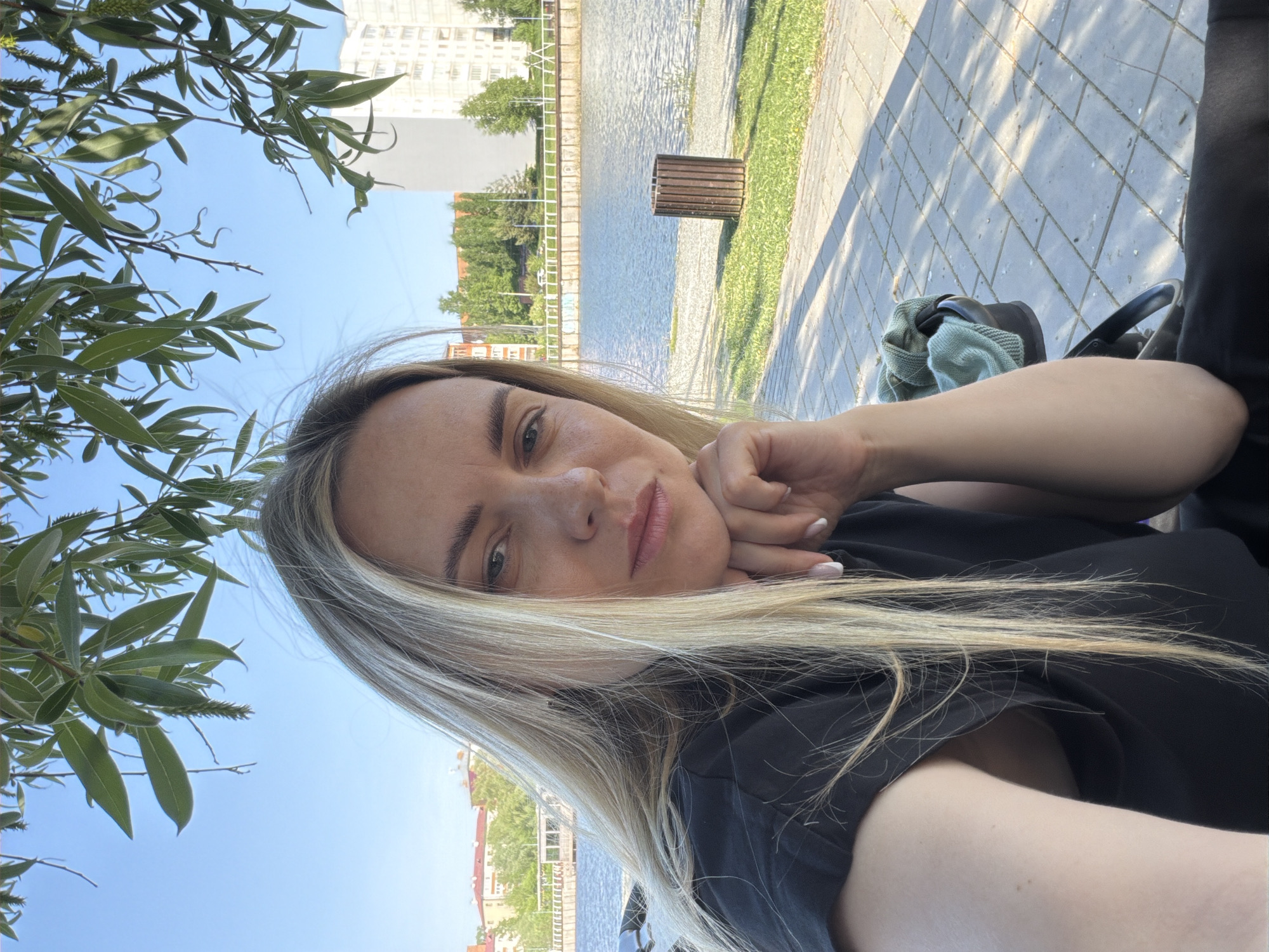 A woman is taking a selfie outdoors. The location appears to be along a waterway, possibly a canal or river, within the city of Votkinsk, Russia. She is leaning her face on her hand. Her hair is blonde and partially obscures her face. She is wearing a dark-colored shirt. In the background, there is a paved walkway, grass, water, and buildings. Tree branches are visible in the upper left of the frame. A stroller and a light green bag are next to the woman.