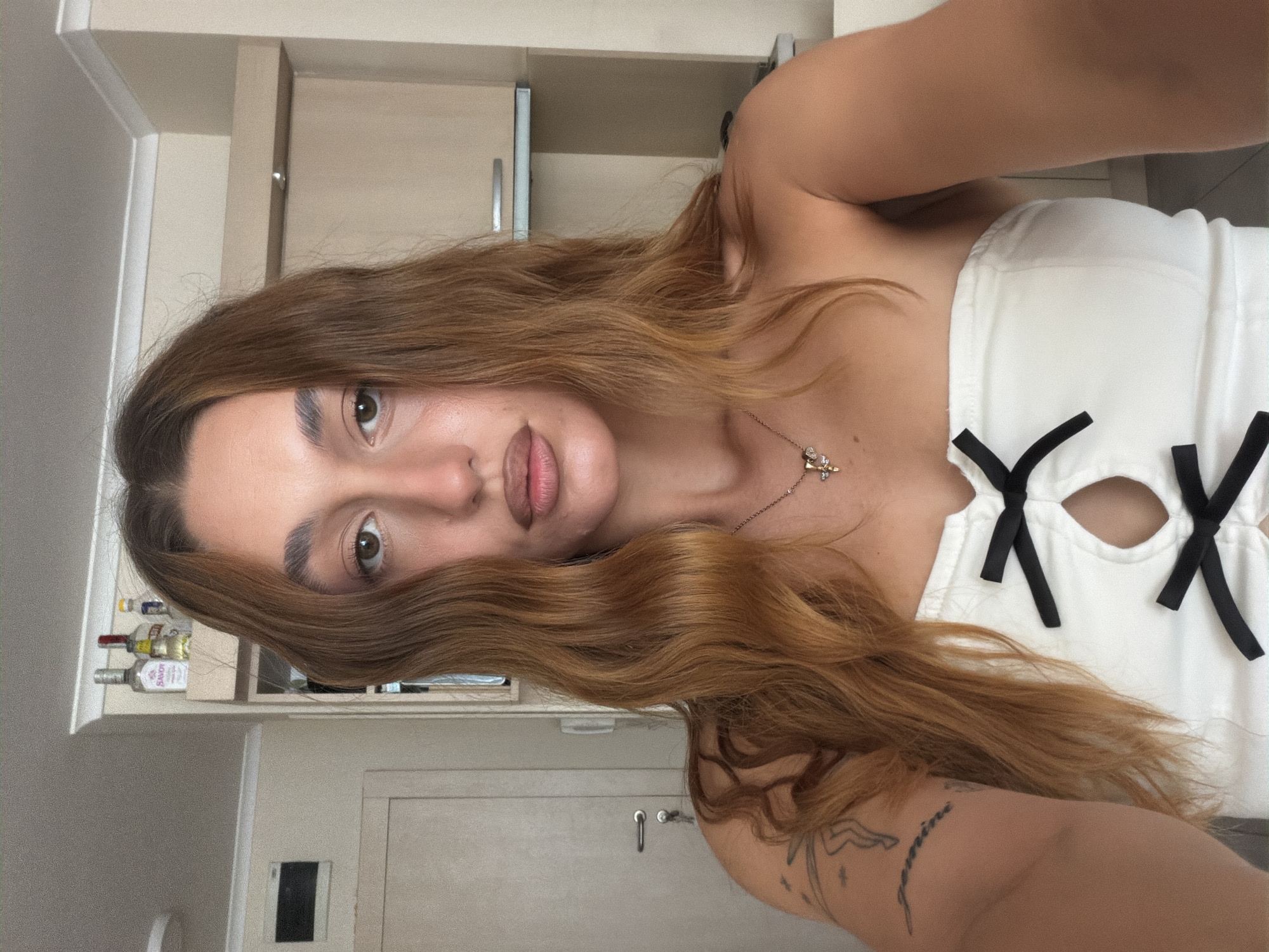 The image is a selfie of a woman with long, wavy brown hair, taken indoors. She is wearing a white strapless top with black ribbon details and a keyhole opening. A necklace with a cross pendant is visible. She has a tattoo on her left arm. The background shows a kitchen area with cabinets and shelves holding bottles, and a door to the left. The lighting suggests daytime. The woman is looking directly at the camera. The image does not provide information about the city of Sofia, Bulgaria, or any specific location within it.