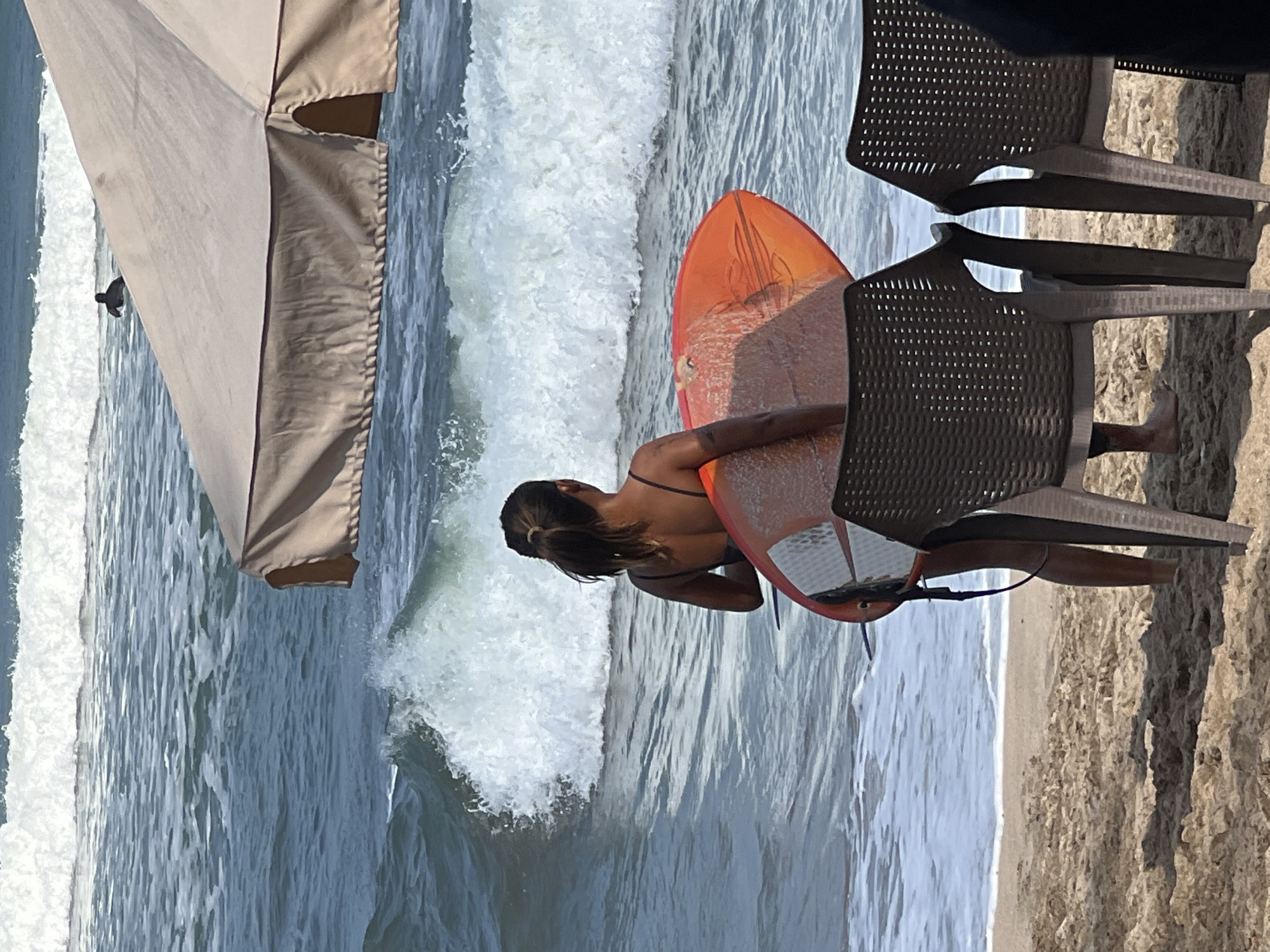 An overcast, daytime scene depicts a sandy beach area in Kuta, Indonesia, where a young woman is holding a vibrant orange surfboard. She stands with her back to the viewer, facing the ocean. Her dark hair is pulled back into a ponytail. She wears a dark-colored swimsuit. In the background, large waves crash against the shore, with white foam spreading across the water. A large, light-colored beach umbrella provides shade in the middle ground. Two dark-colored, woven chairs are positioned in the foreground, obscuring part of the woman's legs. Another individual can be seen further out in the ocean, also with a surfboard. The overall atmosphere suggests a leisurely day at the beach with opportunities for surfing.