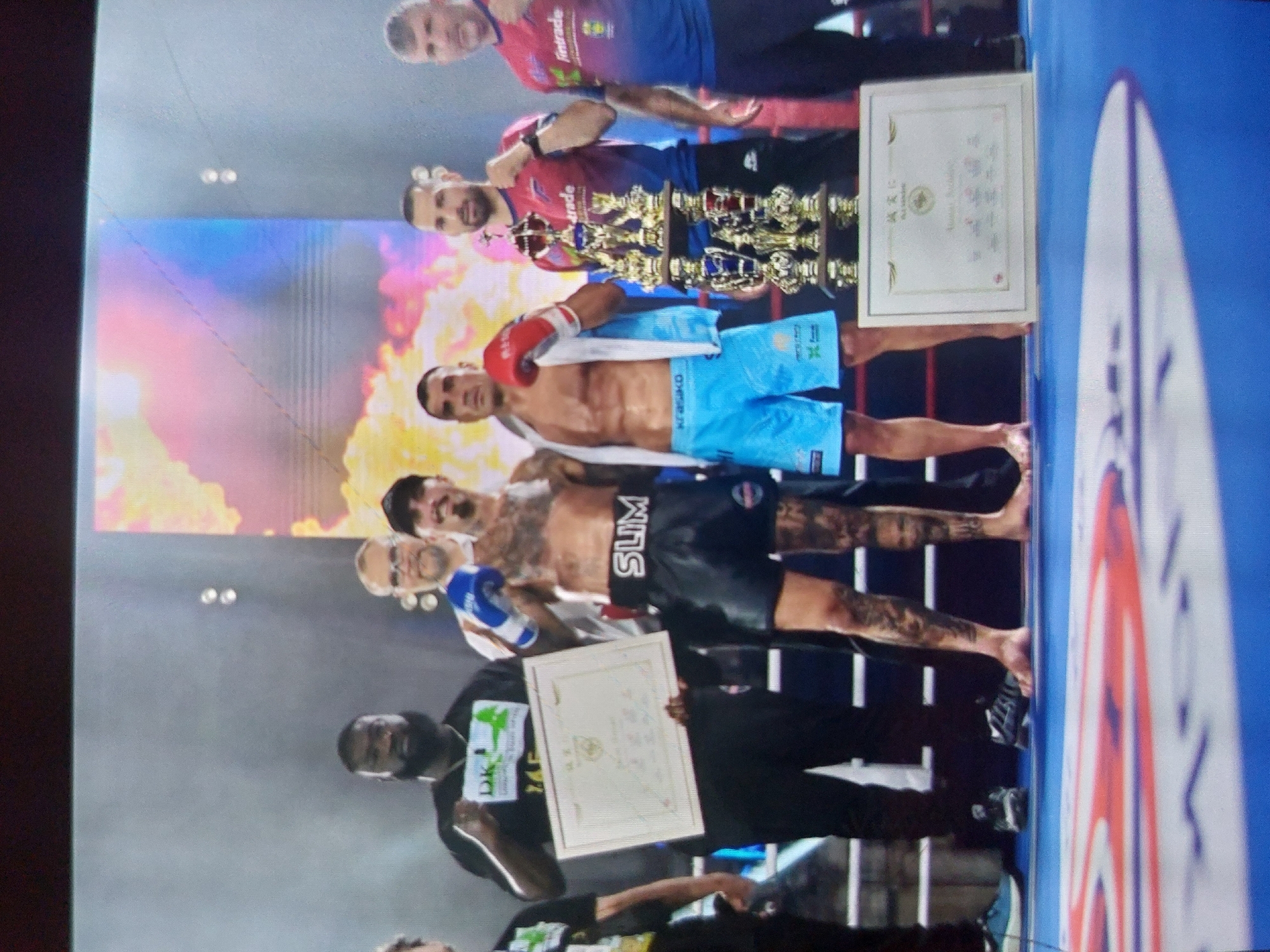 This vertically oriented image captures an awards ceremony following a combat sports event, likely in Tsarevo, Bulgaria. The scene is set on a brightly lit stage or platform, featuring a dynamic backdrop with fiery orange, yellow, red, and blue abstract patterns.
Approximately ten adult men are visible, comprising athletes and officials. In the foreground, two shirtless men, presumably competitors, stand prominently. The man on the left has extensive tattoos on his arms and legs, wears black shorts with "SLIM" written on them, and a blue boxing glove on his left hand. He holds a framed white certificate with a gold border. Slightly behind him and to the right, another shirtless man wears light blue shorts marked "KNECKO" and a red boxing glove on his left hand.
Several other men are present, some holding a large collection of golden trophies of various sizes. Two men at the top appear to be presenting these awards; one wears a blue and red top with "Intrade" visible. Another man in a black t-shirt at the bottom left also holds a framed white certificate similar to the one held by the tattooed athlete. An older man with glasses is also part of the group.
On the far right, a blue and white barrier, characteristic of a boxing or martial arts ring, is partially visible. The atmosphere is celebratory, with smiles suggesting pride and accomplishment. The certificates feature an official seal and what appears to be Asian script, suggesting the specific martial arts discipline. The event likely took place in the evening or at night, indicated by the artificial stage lighting.