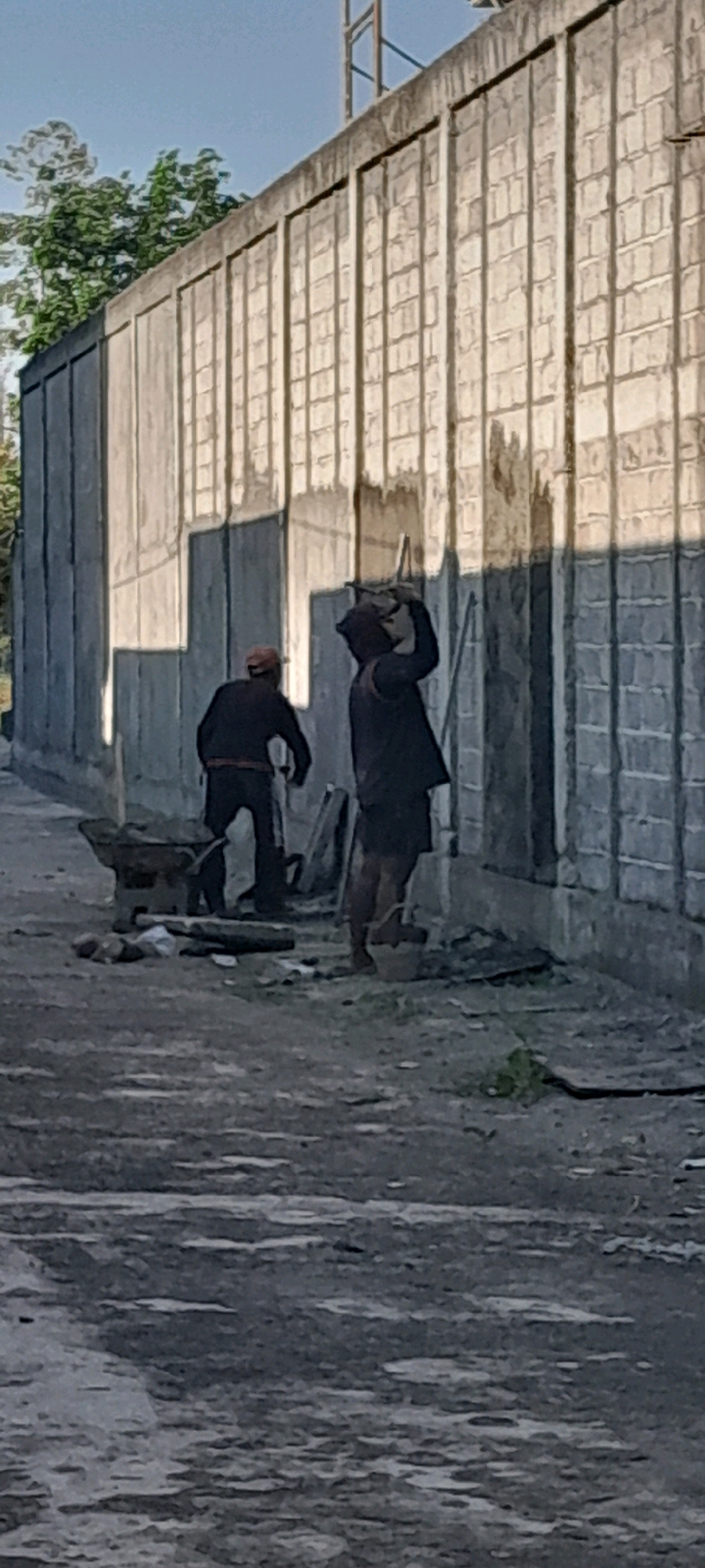 The image depicts an outdoor construction site in Rogojampi, Indonesia, characterized by a long, textured concrete wall running horizontally across the frame. The wall, composed of rectangular blocks, is partially illuminated by sunlight, casting strong shadows. In the foreground, a rough, unpaved ground surface is visible, littered with debris and construction materials.
Two individuals are engaged in work. On the left, a person wearing dark clothing and a red cap stands near a wheelbarrow filled with what appears to be sand or aggregate. Their posture suggests they might be preparing to load or unload materials. On the right, another person in dark clothing, including a hooded garment, is actively working on the wall. They are holding a tool, possibly a trowel or hammer, and are positioned as if applying something to the wall or breaking it down. A bucket or container rests on the ground near their feet.
The environment appears to be a typical urban or semi-urban construction area, with a clear sky visible in the upper portion of the image. The overall scene conveys a sense of ongoing manual labor and construction activity.