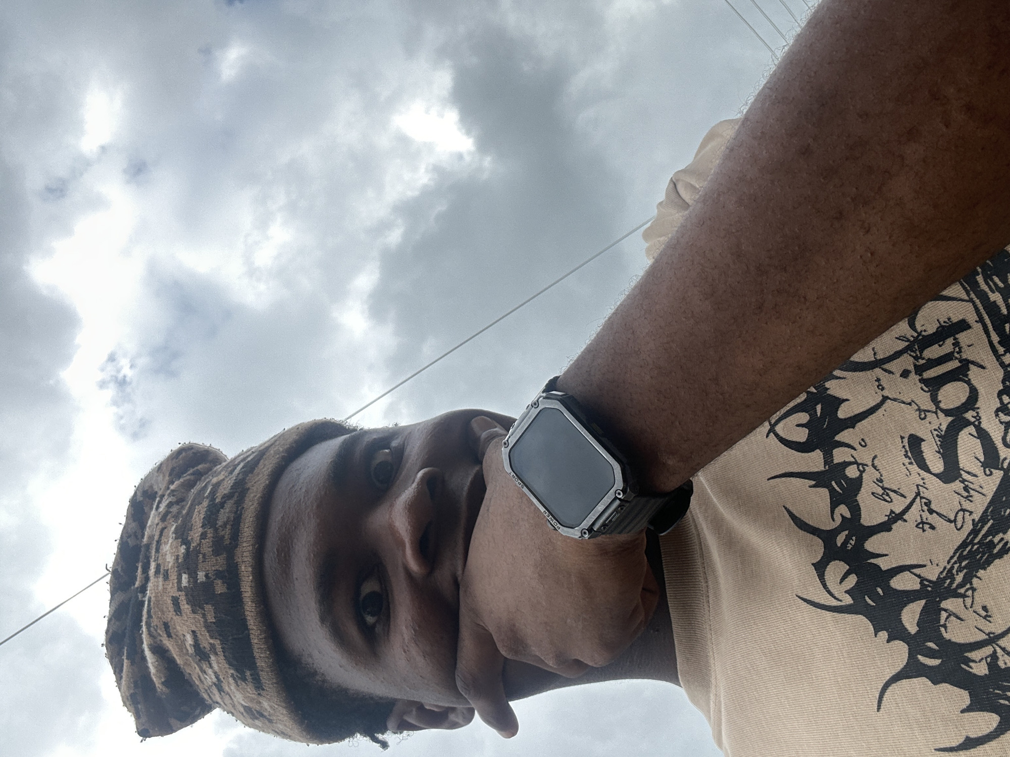 This is a close-up, low-angle selfie taken outdoors, with a cloudy sky as the dominant background. The image is oriented sideways, showing a person from the chest up. The subject is wearing a patterned beanie and a light-colored t-shirt with a bold, black graphic design on it. On the subject's left wrist, a chunky, dark-colored smartwatch with a metallic bezel is visible. The person's right hand is partially covering their mouth and chin, with their eyes looking towards the camera. The weather appears overcast, suggesting it might be daytime under cloudy skies. The visible text on the smartwatch reads "SPORT" and "POWER". The overall mood is casual and somewhat introspective. The context provided places the location as Ihiagwa, Nigeria.