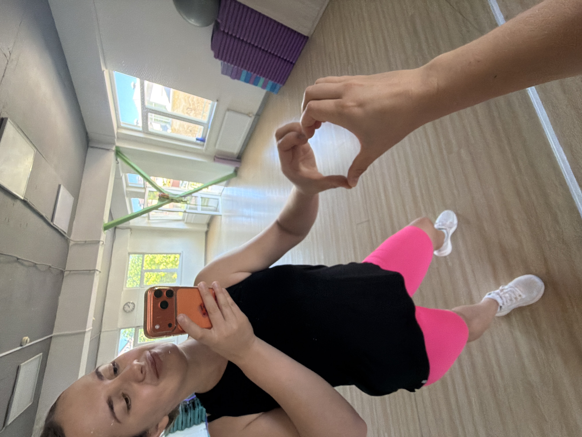 A woman is captured in a mirror selfie within what appears to be a fitness studio or gym in Sochi, Russia. The image is taken from a slightly high angle, reflecting the woman, the room, and some outdoor scenery.
The main subject is a woman with dark hair, wearing a black tank top, bright pink cycling shorts, and white athletic shoes. She holds an orange-cased smartphone in her right hand to take the photo. Her left hand is joined with what appears to be her right hand (partially out of frame) to form a heart shape, positioned prominently in the upper right portion of the reflection. She has a neutral to slightly smiling expression on her face.
The setting is an indoor space with light-colored wooden flooring and light gray walls. In the background, behind the woman, multiple large windows reveal an outdoor scene with green trees and buildings, indicating that it is daytime. Stacked purple and blue exercise mats are visible in one corner. There's also a green pole or beam structure running across the room, possibly part of the building's support or gym equipment. Several white rectangular frames or boards are mounted on one of the gray walls. A small, white circular clock is visible on the wall through one of the windows.
The overall impression is that of a casual moment, perhaps after a workout, where the woman is expressing positivity through the heart gesture. There is no visible text in the image.