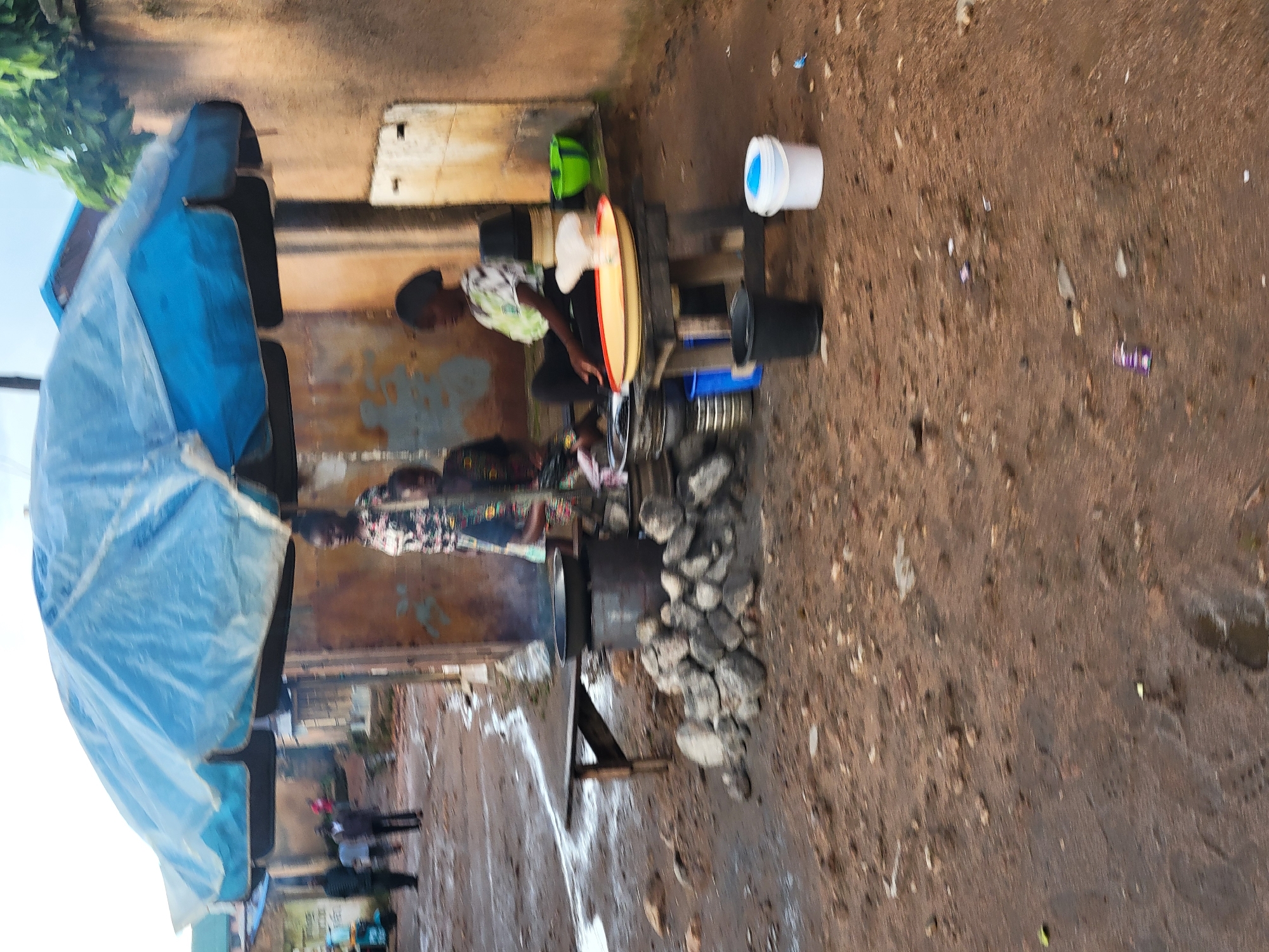 A group of people are gathered around an outdoor food stall under a large blue umbrella. The stall appears to be in a village or market setting with a dirt ground. There are several cooking pots and utensils visible, suggesting food preparation is taking place. Some individuals are standing and observing, while others are seated. The overall scene depicts a typical day at a local food vendor in a community.