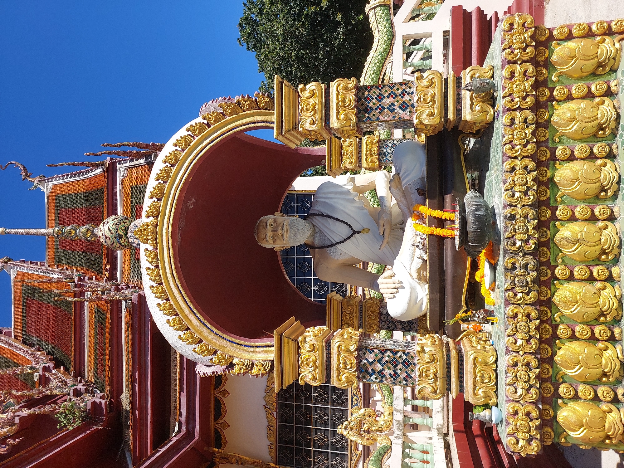A statue of an elderly man with a white beard and robes is seated in a shrine at the Wat Plai Laem temple in Baan Plai Laem, Thailand. The statue is made of white material and has a black beaded necklace. The shrine is ornate with gold trim and colorful tiled pillars. Yellow and orange flower garlands are placed in front of the statue. The temple's tiled roof is visible in the background under a clear blue sky. Lush green trees are also present in the background.