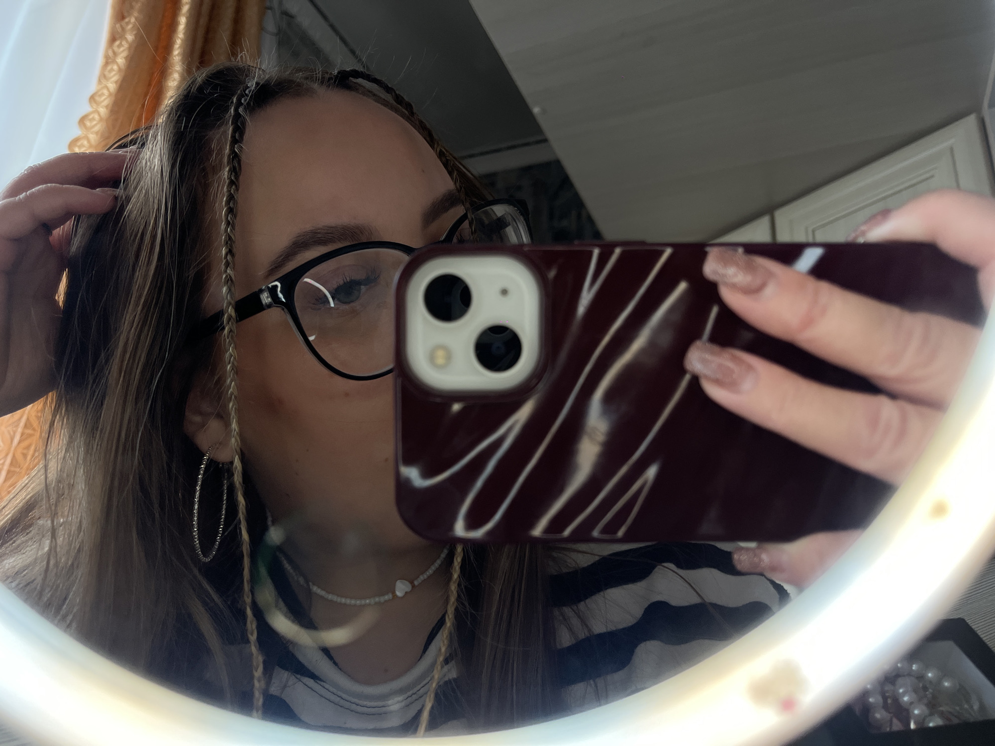 A woman is taking a selfie in a mirror. She has long brown hair with small braids near her face and is wearing glasses, hoop earrings, and a white beaded necklace with a heart charm. She is holding a maroon-colored smartphone in front of her face. A white ring light surrounds the mirror. The background includes a striped shirt and what appears to be a room with curtains. The exact location is not discernible as being in Novopokrovskaya, Russia, from the provided image.