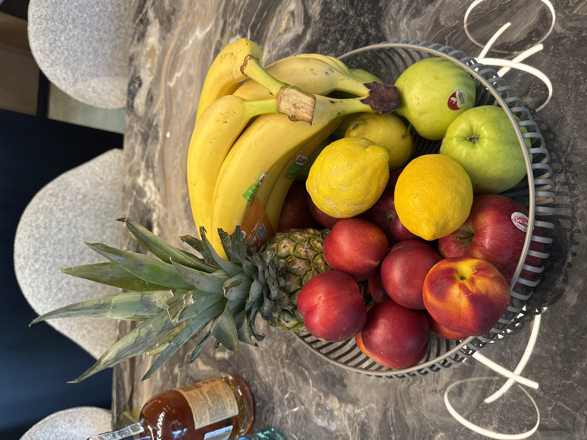 A close-up shot presents a woven wire fruit basket filled with a vibrant assortment of fresh produce. Bright yellow bananas and lemons are interspersed with red apples and peaches. Green guavas add to the variety, and a pineapple with its characteristic green spiky leaves is positioned at the edge of the basket. The basket is placed on a mottled grey countertop. A glass bottle with a brown liquid and a blue object are also visible near the basket. The location appears to be indoors.
