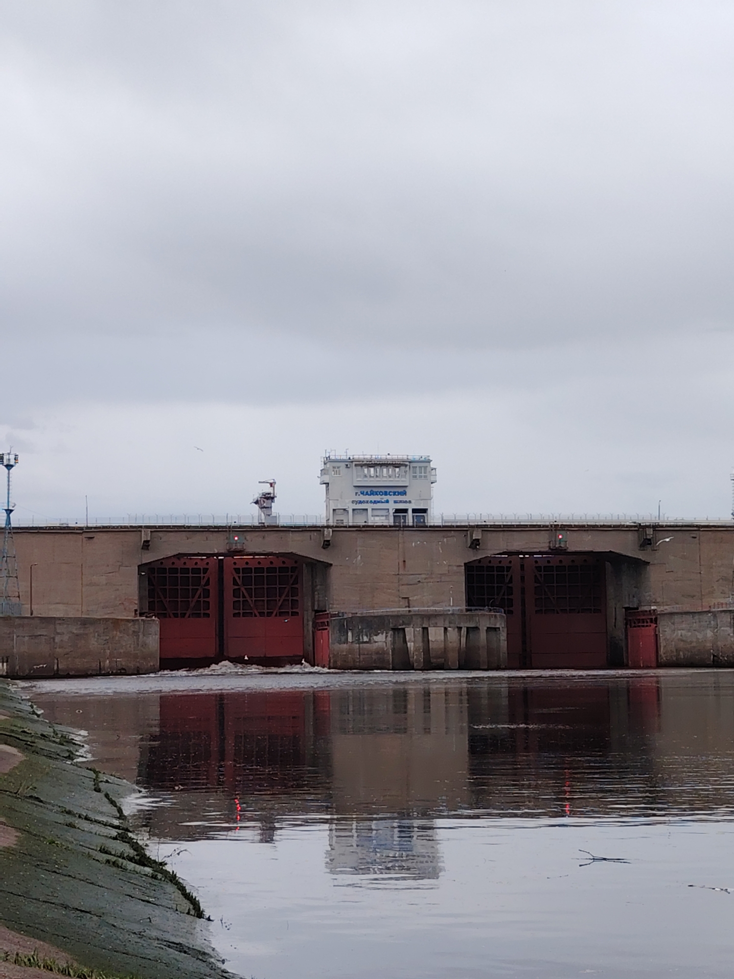 The image shows the Shlyuzy Chaikovskoy GES, which are the locks of the Chaikovsky Hydroelectric Station, located in the city of Chaikovsky, Russia. The gray sky is overcast. The river is in the foreground. The lock structure consists of reinforced concrete with large red steel gates. The water level behind the lock appears higher than the water level in front of the lock, indicating the lock is open and releasing water. Reflections of the structure are visible in the water. There's greenery along the edge of the water in the foreground.