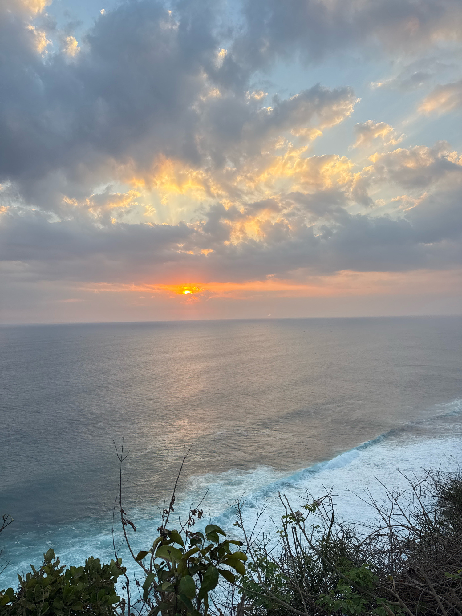 The image depicts a sunset over a vast ocean, viewed from a cliffside. The sky is filled with dramatic clouds, tinged with orange and yellow from the setting sun, which is partially obscured by clouds and appears as a bright orb on the horizon. Rays of light pierce through the clouds, casting a warm glow on the scene. The ocean is a deep blue, with white waves crashing against the shore at the bottom right of the frame. The foreground is dominated by dark, dry vegetation, characteristic of a cliffside environment. The overall impression is one of natural beauty and tranquility. The location is not specified in the city of Unknown, Unknown.