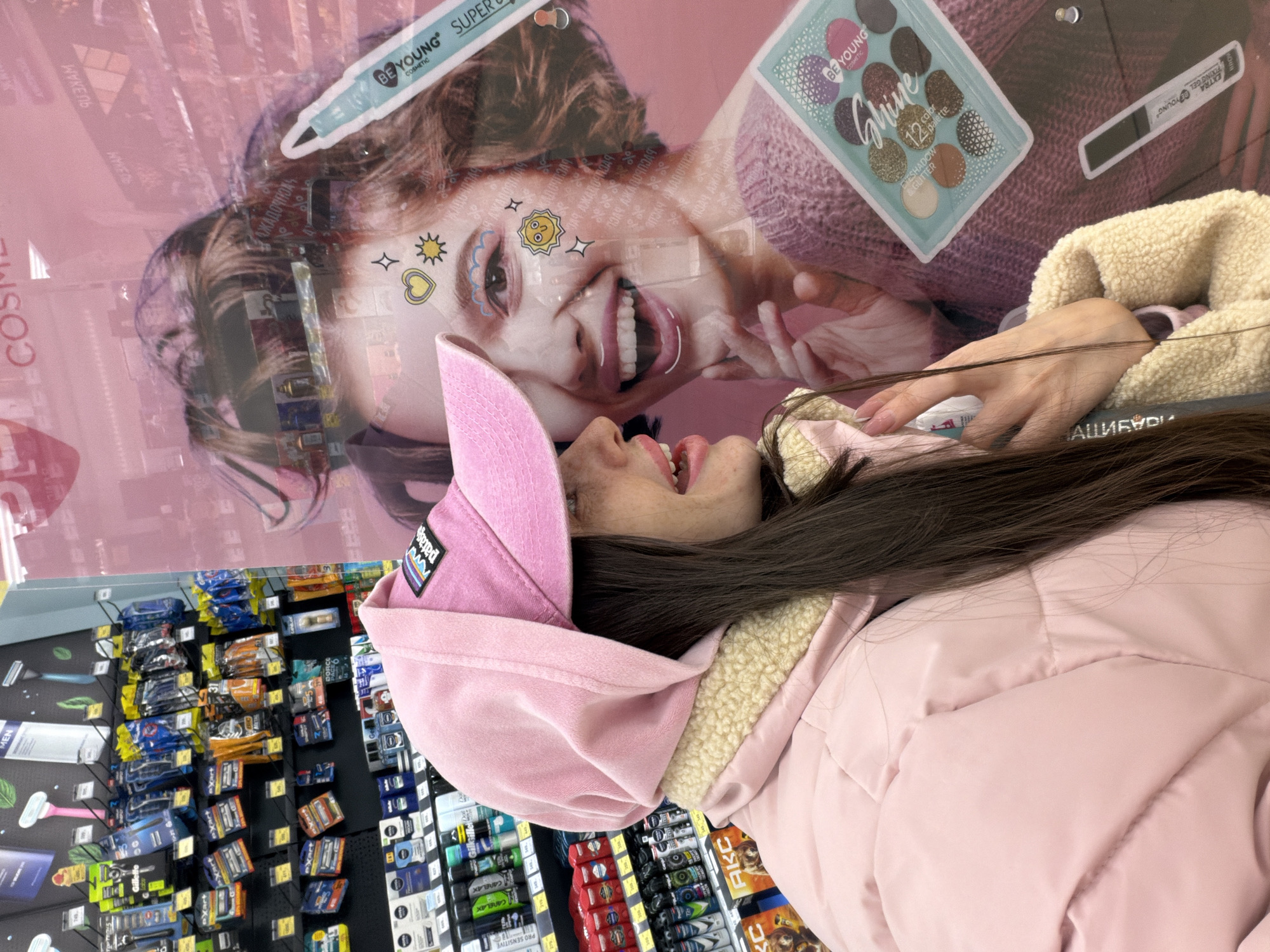 A female individual is captured from the mid-torso up, standing inside what appears to be a brightly lit retail store in Saint Petersburg, Russia, likely a supermarket or hypermarket. She is positioned facing right in profile, looking towards her left, seemingly interacting with or observing a large, reflective store display or glass wall featuring a pink-themed cosmetic advertisement.
The individual wears a light pink padded jacket with a cream-colored sherpa fleece lining visible around the collar and hood. Her hood is up, and she wears a light pink baseball cap with a dark, rectangular "Patagonia" logo patch on the front, over her long, dark brown hair which cascades over her shoulder. Her mouth is slightly open, revealing white teeth, and her lips appear to have a glossy pink tint. Her right hand is partially visible near her chin, with neatly manicured fingernails.
The background to the left of the individual reveals multiple shelves stocked with various personal care products, including razors (e.g., Gillette), deodorants (e.g., Nivea, Gillette, Care & Clean), and other toiletries. Product prices are visible on yellow labels. A magazine or display with the Russian text "АКС" (AKS) is partially visible at the bottom left.
The reflective surface or transparent poster on the right features a large image of a smiling woman with light brown, curly hair, adorned with whimsical facial stickers (sun, heart, stars, cloud shapes). This model is advertising cosmetics, including a "BE YOUNG" brand eyeshadow palette labeled "Shine 12 color eyeshadow & glitter palette" and a "BE YOUNG Super Gel" pen. The reflection of additional store shelves with cosmetic products and overhead lighting is visible superimposed over the model's image. Russian text such as "КОСМЕТИКА" (Cosmetics) and "МАКИЯЖ" (Makeup) is faintly visible on the advertising.