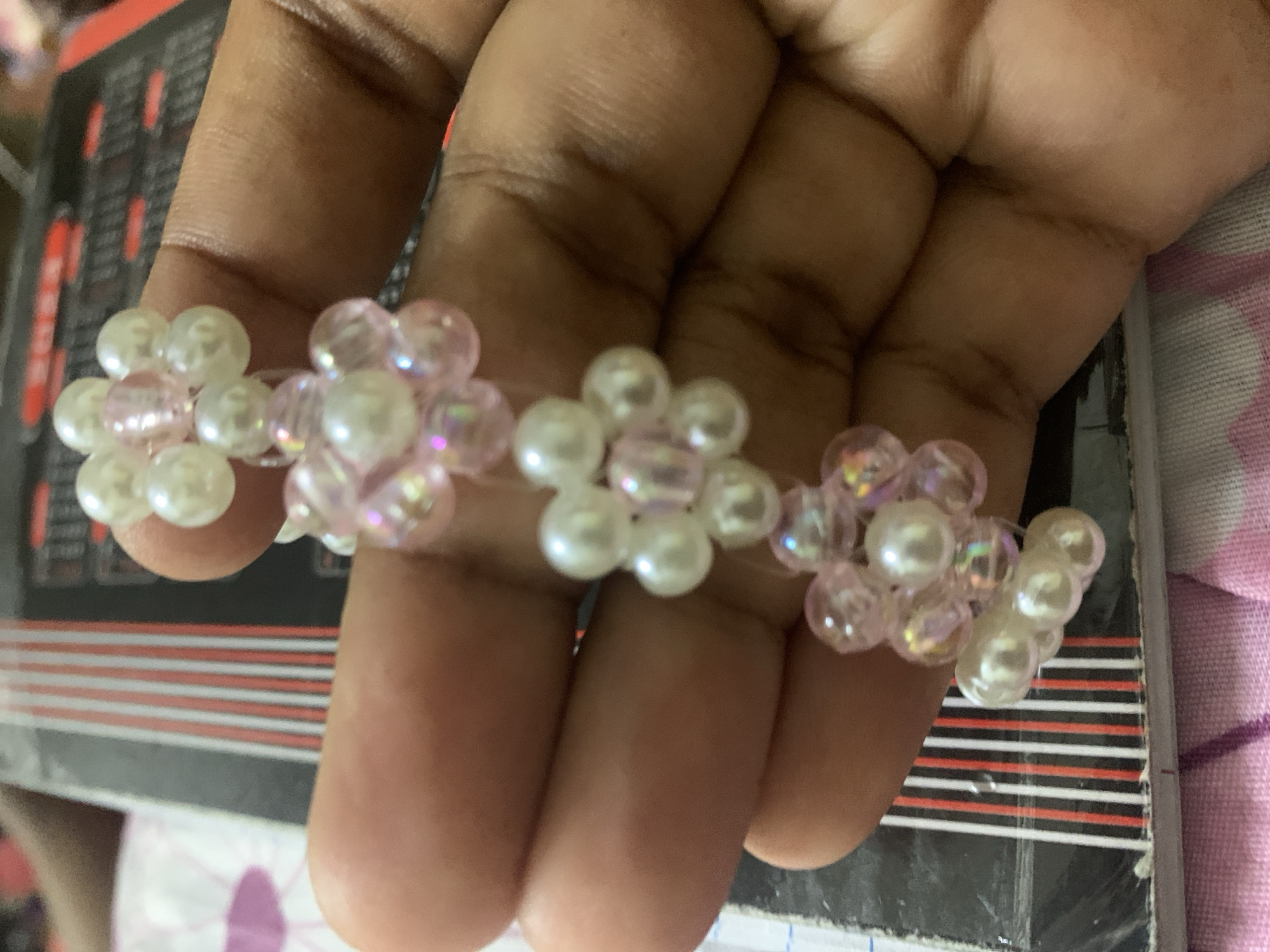 A hand holds a beaded bracelet with alternating pink and white beaded flower designs. The background shows a book with red and white stripes, and part of a pink patterned surface. There is no discernible information to suggest the location is Dar es-Salaam, Tanzania.