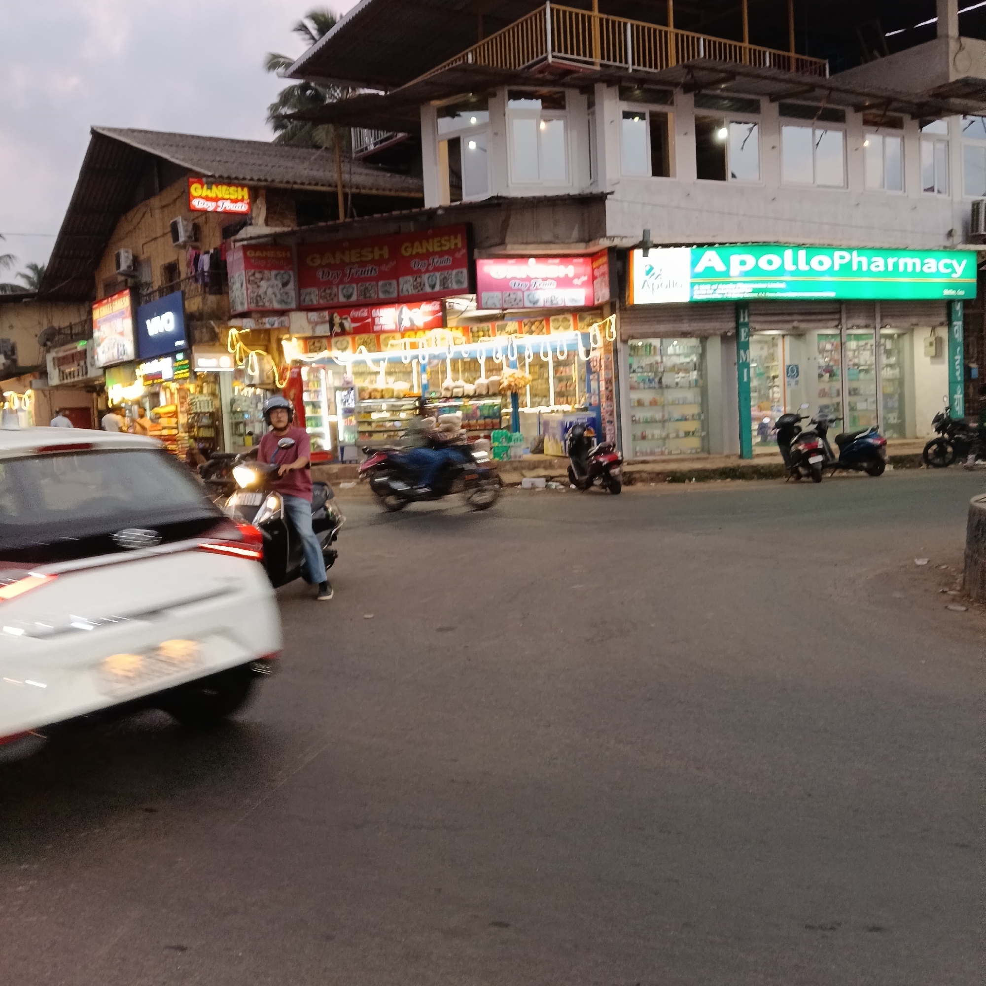 The image depicts a street scene in Oxel, India, featuring a variety of establishments. The Apollo Pharmacy is visible on the right side of the image. Adjacent to it are shops such as "GANESH Dry Fruits", which appear to be a sweets and snack retailer. There's also a "Vivo" sign, indicating an electronics store. There are several scooters parked along the sidewalk. A person on a scooter is visible in the foreground near a white car in motion. The street appears to be asphalt. The buildings are of various heights with a mix of traditional and modern architectural styles, some with multiple stories and balconies. The overall atmosphere suggests a bustling commercial area.