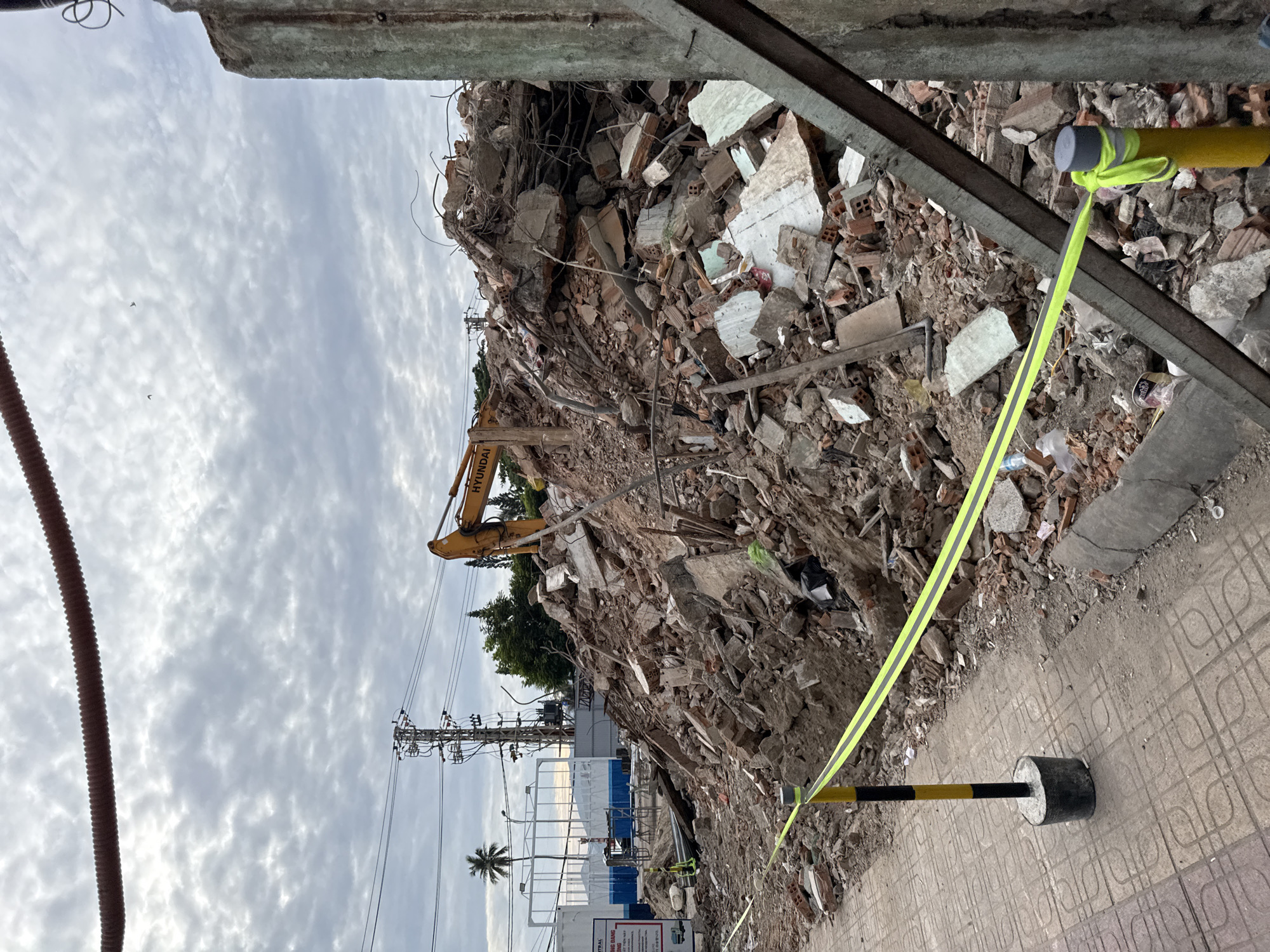 The image depicts a construction demolition site in Nha Trang, Vietnam. A large pile of rubble, consisting of broken concrete, bricks, and rebar, dominates the center of the frame. A yellow Hyundai excavator is partially visible at the top of the debris pile. In the foreground, a yellow and black striped pole supports a bright yellow caution tape that runs across the scene, separating the construction area from a tiled pedestrian walkway. The walkway is bordered by a concrete curb. In the background, power lines and a palm tree are visible against a cloudy sky. A large corrugated pipe runs horizontally across the top of the image. A concrete pillar is visible on the right side of the frame. There are no people clearly depicted in the scene, though it is possible some are present within the rubble or out of frame.