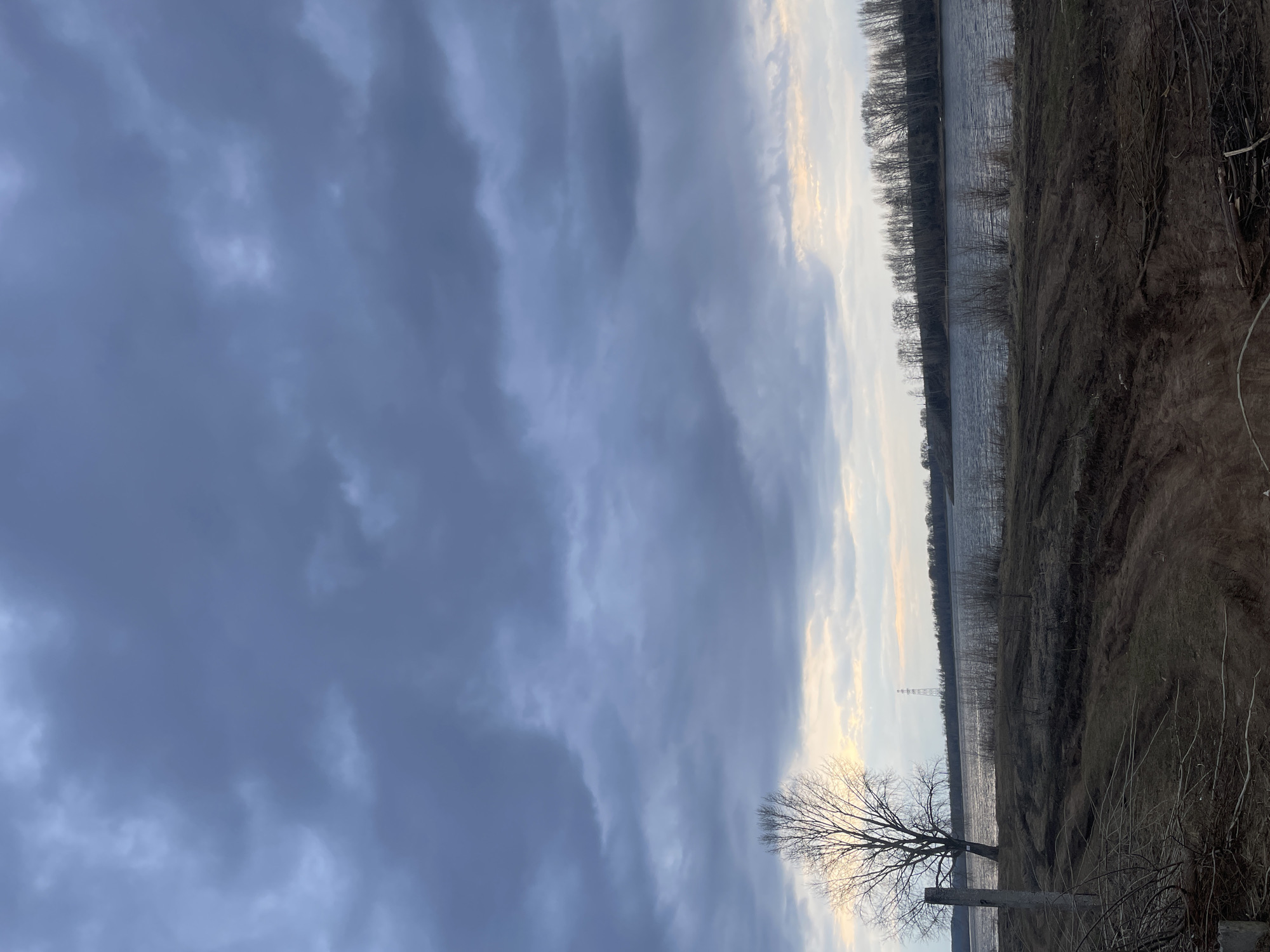 The image depicts a landscape with a body of water, likely a river or lake, on the right side. Along the shore, there's a strip of trees and land. The sky is predominantly filled with dark, heavy clouds. In the foreground, a bare tree stands near a concrete or stone structure. In the distance, there's a power line tower visible on the horizon. The scene appears to be taken in a natural environment with a cloudy sky. The exact location in Chaikovsky, Russia, is impossible to determine from this single image.