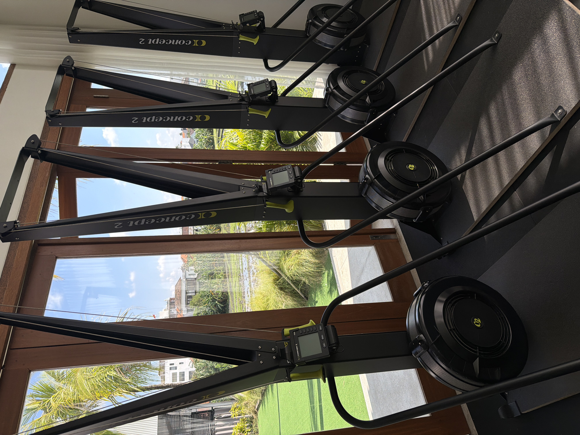 This image captures a row of four black Concept 2 rowing machines, positioned indoors within what appears to be a gym or fitness area. The machines are uniformly arranged, facing outward through large windows with dark wooden frames. Each rower features a black frame, a monitor displaying "Concept 2" (visible inverted on some), a black flywheel with a distinct green "C2" logo, and yellow handles. They rest on a dark, textured floor, likely rubber matting.
Beyond the windows, a bright, sunny outdoor scene is visible. The sky is clear blue with scattered white clouds. Lush green tropical foliage, including palm trees, dominates the immediate outdoor view, hinting at a warm climate. In the distance, several buildings with light-colored facades and various roof styles can be seen. Through the lower windows, a vibrant green area resembling a manicured lawn or sports court is prominent. No people are present, but the setup clearly indicates a space designated for exercise. The tropical vegetation and residential architecture align with the provided location context of North Kuta, Indonesia.