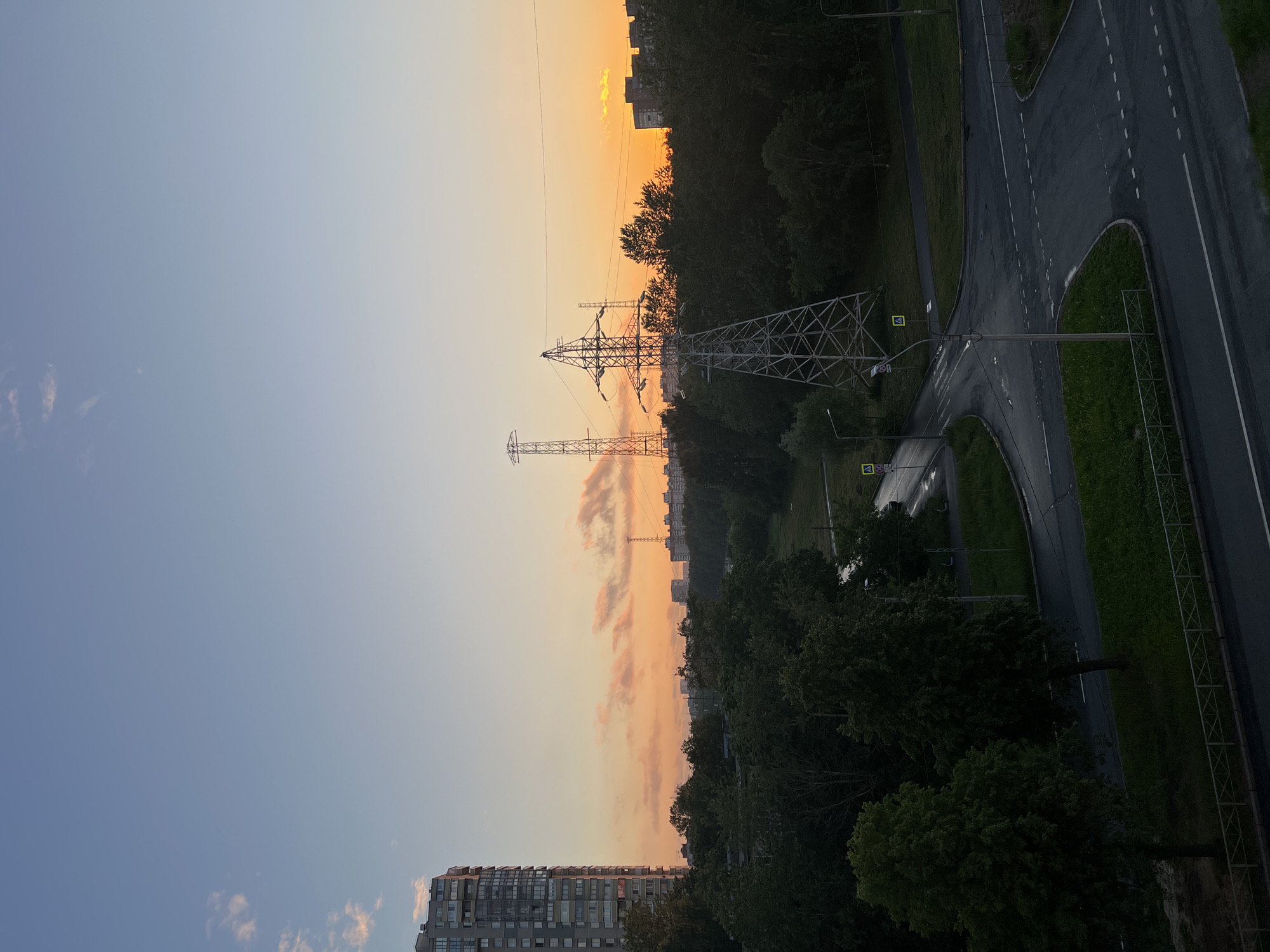 This is an aerial, rotated landscape photograph taken at dusk in Saint Petersburg, Russia. The scene captures a transition from day to night, with a vibrant orange and yellow sunset illuminating the sky on the left side of the frame, fading into a soft blue on the right. A large, modern apartment building with many balconies is visible in the lower left.
Dominating the mid-ground are two towering metal electricity pylons, silhouetted against the colorful sunset. Below the pylons and stretching across the right side of the image is a wet, asphalt road, likely from recent rain, with clear white lane markings. The road curves through a patch of lush green grass. Tall, dark green trees line the edge of the road and fill the rest of the right and bottom portions of the frame. In the distance, more apartment buildings and trees are visible, suggesting an urban or suburban setting.
Several road signs are visible, including a yellow triangular sign with a black symbol and rectangular signs with red and white text. The overall mood is serene and peaceful, characteristic of a quiet evening in a city.