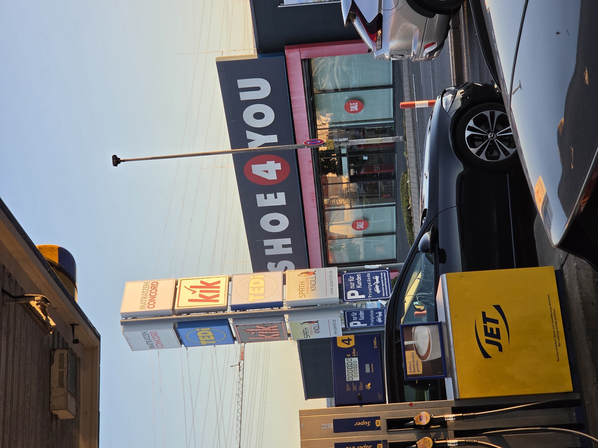 The image depicts a street scene in Bischofsheim, Germany, likely at a gas station and retail area. In the foreground, a yellow JET gas pump is visible, with fuel hoses attached. Next to it, a black car is parked. Behind the car and pump, a row of storefronts with prominent signage is present. One building displays the large white text "SHOE 4 YOU" with a red circle containing a "4" in the center. Red "SALE" signs are affixed to the windows of this establishment. To the left of "SHOE 4 YOU," a tall sign tower displays logos for various businesses, including "MATRATZEN CONCORD," "TEDI," and "kik." Overhead, power lines crisscross the sky. A street lamp stands tall to the right of the sign tower. A silver SUV is partially visible to the right of the "SHOE 4 YOU" building. The overall scene is captured under a bright, possibly late afternoon or early morning sky.