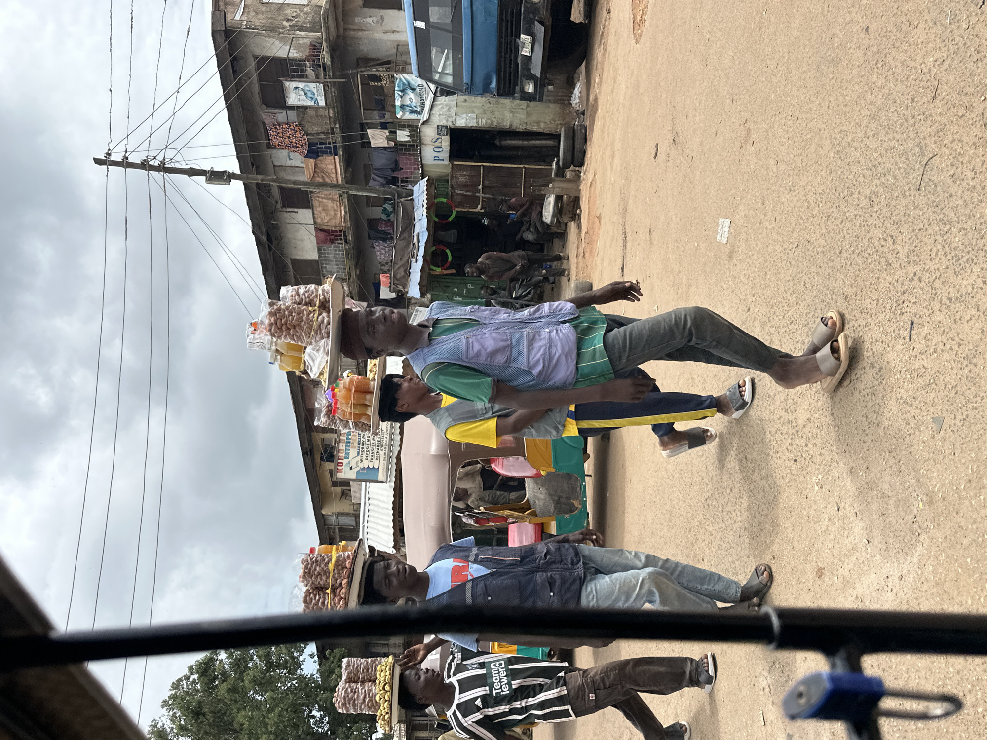 In Jos, Nigeria, a group of young men walk along a dusty street, each balancing a tray of goods on their head. The man in the foreground, wearing a blue vest over a green and white striped shirt and grey pants, walks with his head slightly tilted forward. To his left, a man in a black and white striped t-shirt and brown pants walks briskly, his right hand raised in a gesture of greeting or acknowledgment. Behind them, another young man in a blue t-shirt and dark vest walks, his eyes directed forward. Further back, a man in a yellow shirt and blue pants walks, carrying a tray of items.
The street is lined with buildings exhibiting signs of wear and tear, with balconies adorned with drying laundry. A blue truck is parked on the right side of the street, and a multi-colored auto-rickshaw is visible between the walking figures. The sky above is overcast, suggesting a cloudy day. The ground is a light brown, unpaved surface with scattered debris. The overall scene depicts a vibrant street market atmosphere in Jos.