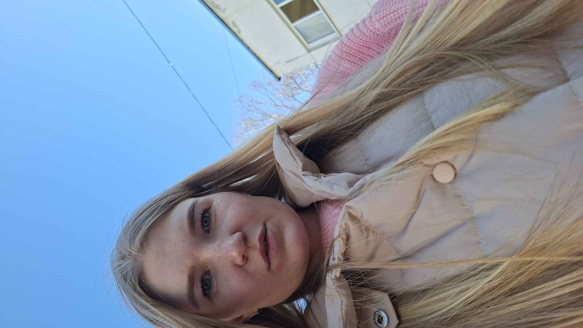 The image is a low-angle selfie of a young woman taken outdoors in Chaikovsky, Russia. The subject is a young woman with long, straight blonde hair, light-colored eyes, and a fair complexion. She is wearing a pink knitted sweater under a light beige or cream-colored puffy jacket. She looks directly into the camera with a neutral expression.
The background consists primarily of a clear, bright blue sky, indicating a sunny day. In the upper portion of the frame, a segment of an older, light-colored building with a window is visible. Bare tree branches can also be seen against the sky, suggesting the photo was taken during winter or early spring. A thin wire or power line crosses the blue sky. The overall scene depicts a casual outdoor setting. No visible text is present in the image.