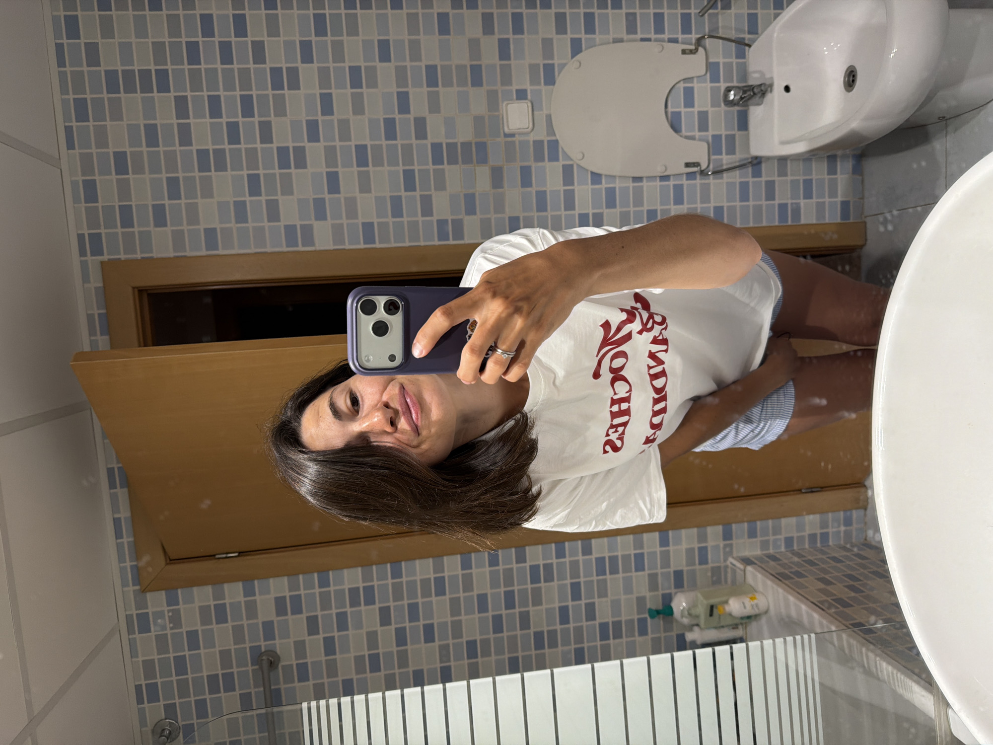 In a bathroom, a woman with dark hair, wearing a white t-shirt and striped shorts, takes a mirror selfie with her smartphone. The bathroom features blue and white checkered tiles on the walls and a wooden door. A bidet is visible in the background, along with a sink and various toiletries on a shelf. The overall scene suggests a casual, domestic setting.